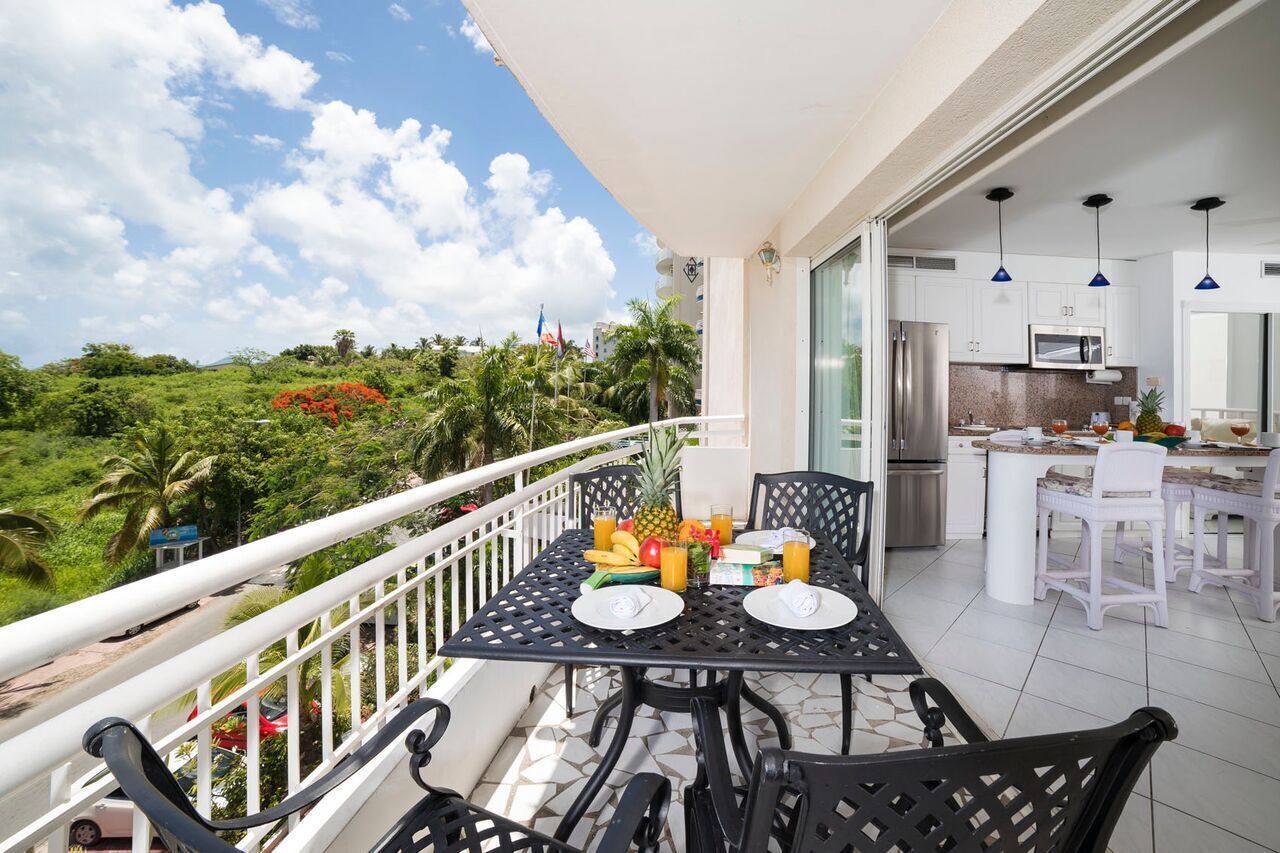 A balcony with a table set for breakfast, a kitchen area, and a view of the surrounding greenery.