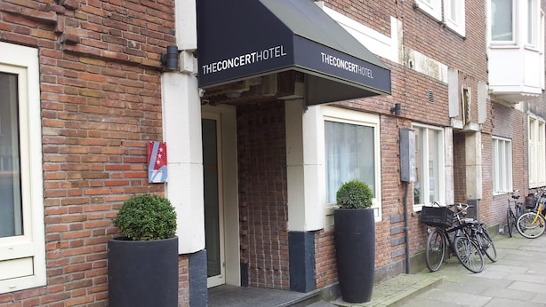 A brick building with a black awning labeled "The Concert Hotel." There are steps leading to the entrance, two potted plants, and bicycles parked outside. - The Concert Hotel (Amsterdam)