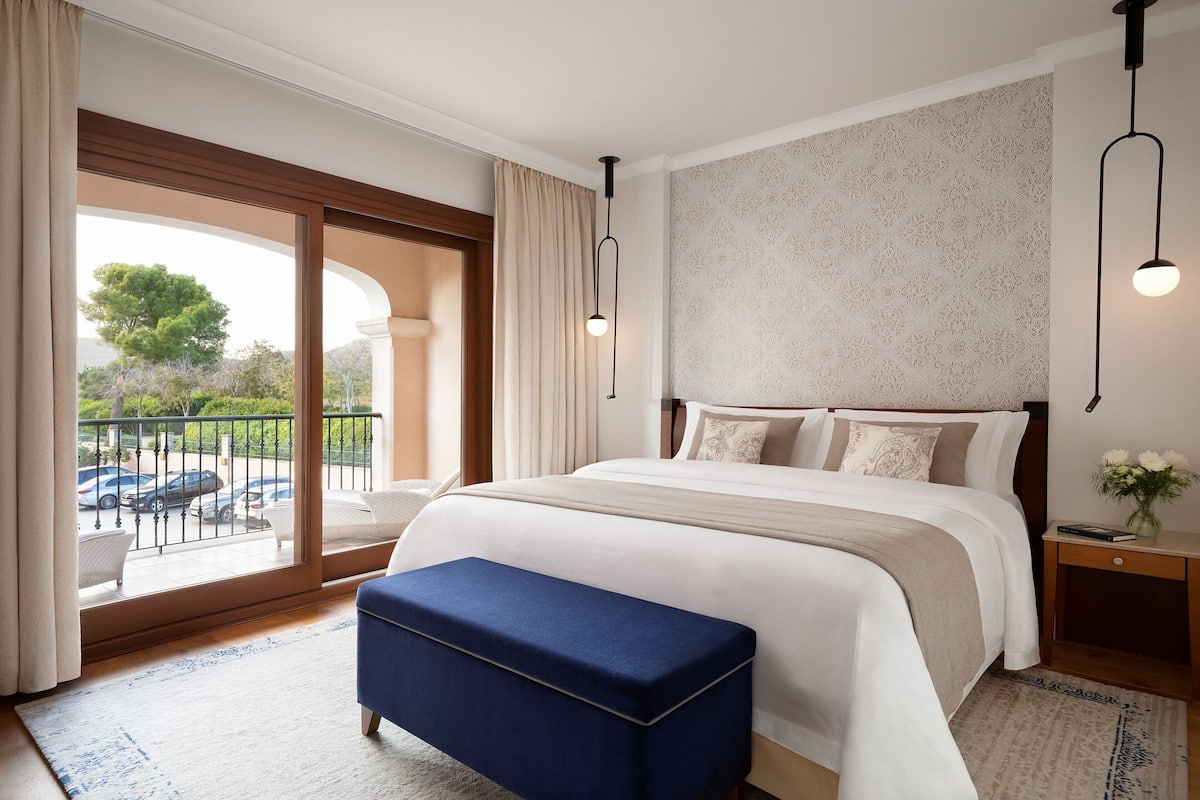 A bedroom with a large bed, a bench, a nightstand, and a view of the outdoors.