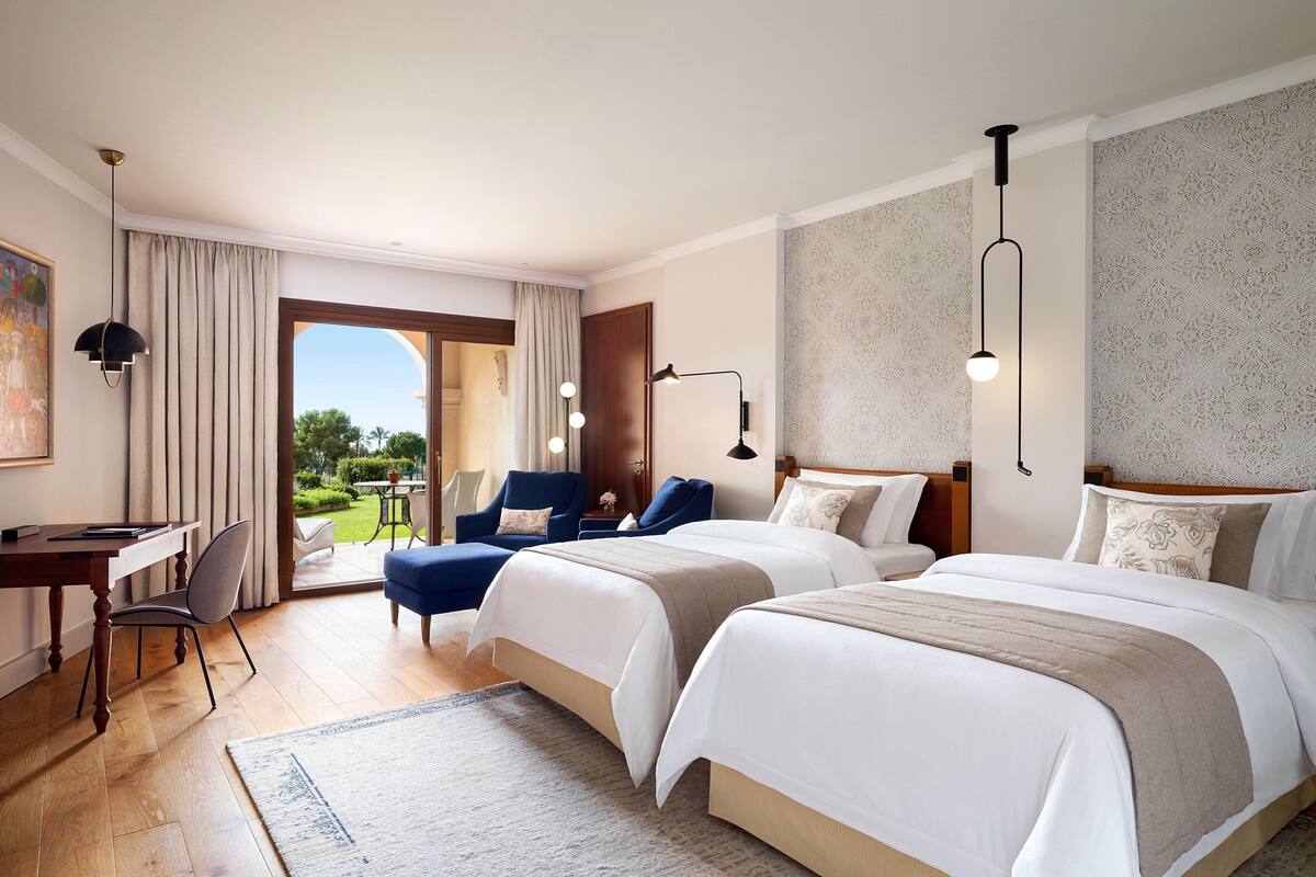 A hotel room with two beds, a desk, a chair, and a view of the outdoors.