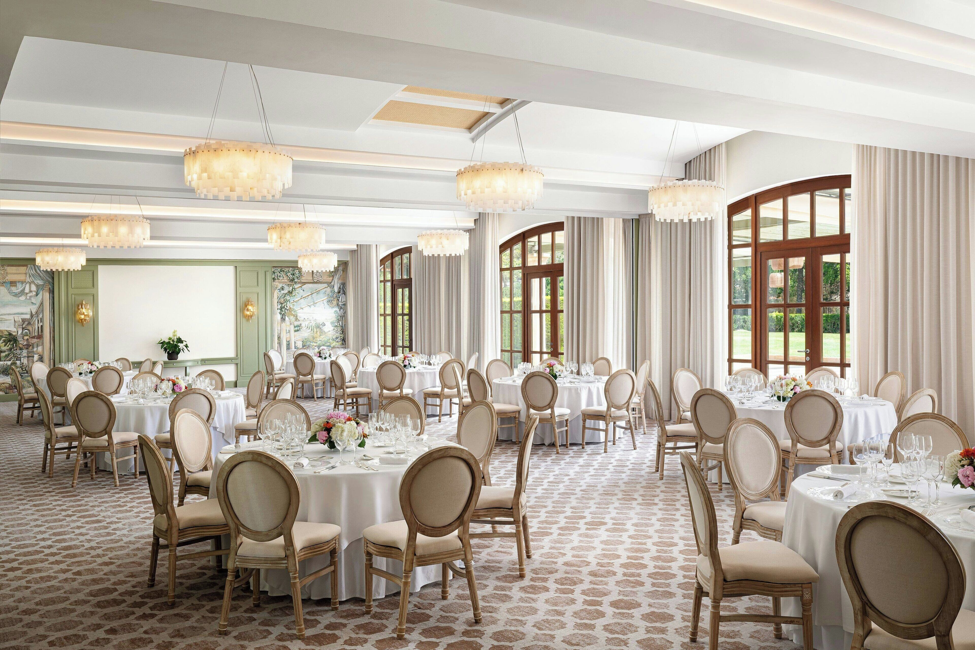 Un salón de banquetes con mesas redondas, sillas, candelabros y ventanas amplias.