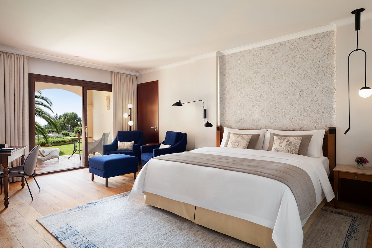 A modern hotel room with a large bed, two blue armchairs, a desk, and a view of the outdoors.