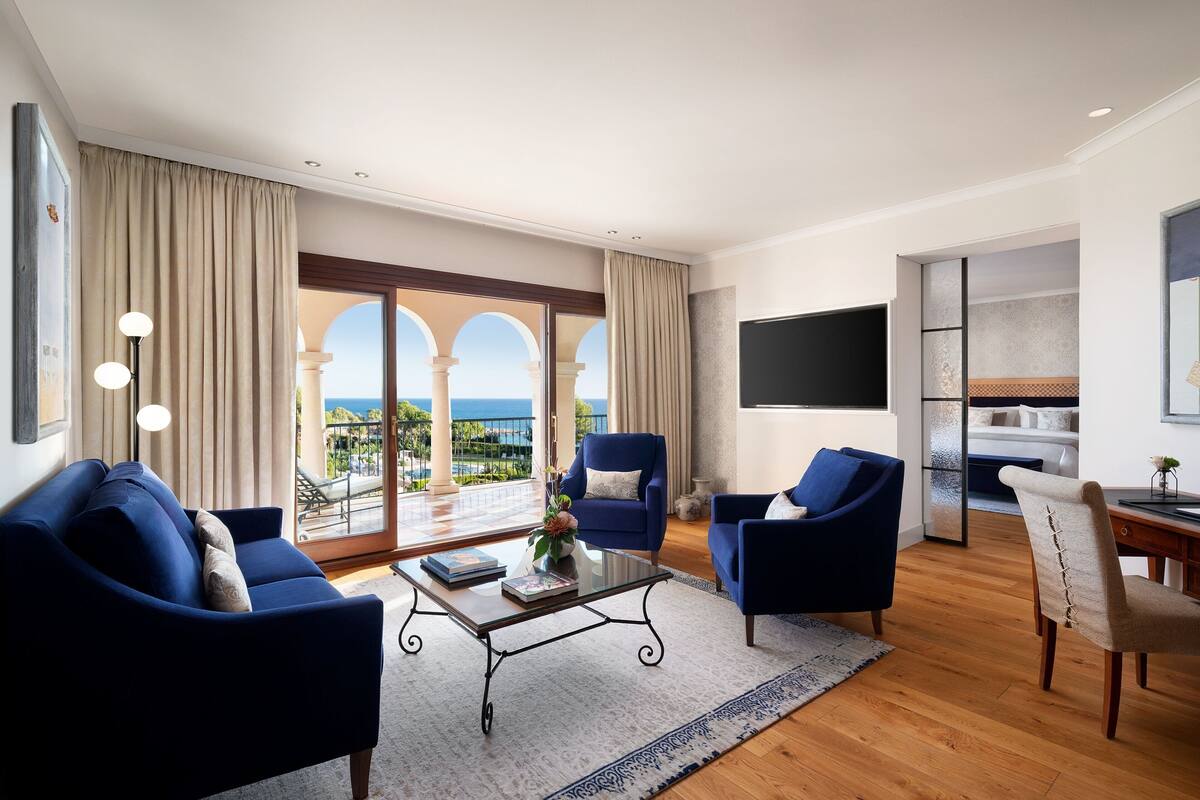 A modern living room with a large sliding glass door leading to a balcony with a view of the sea. There are two blue armchairs, a coffee table, and a TV mounted on the wall.