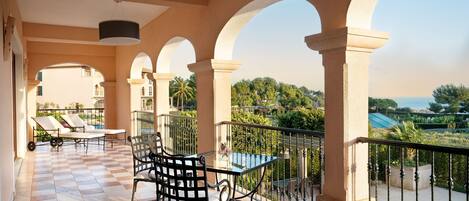 A balcony with checkered flooring, a table, and chairs, overlooking a pool and greenery.