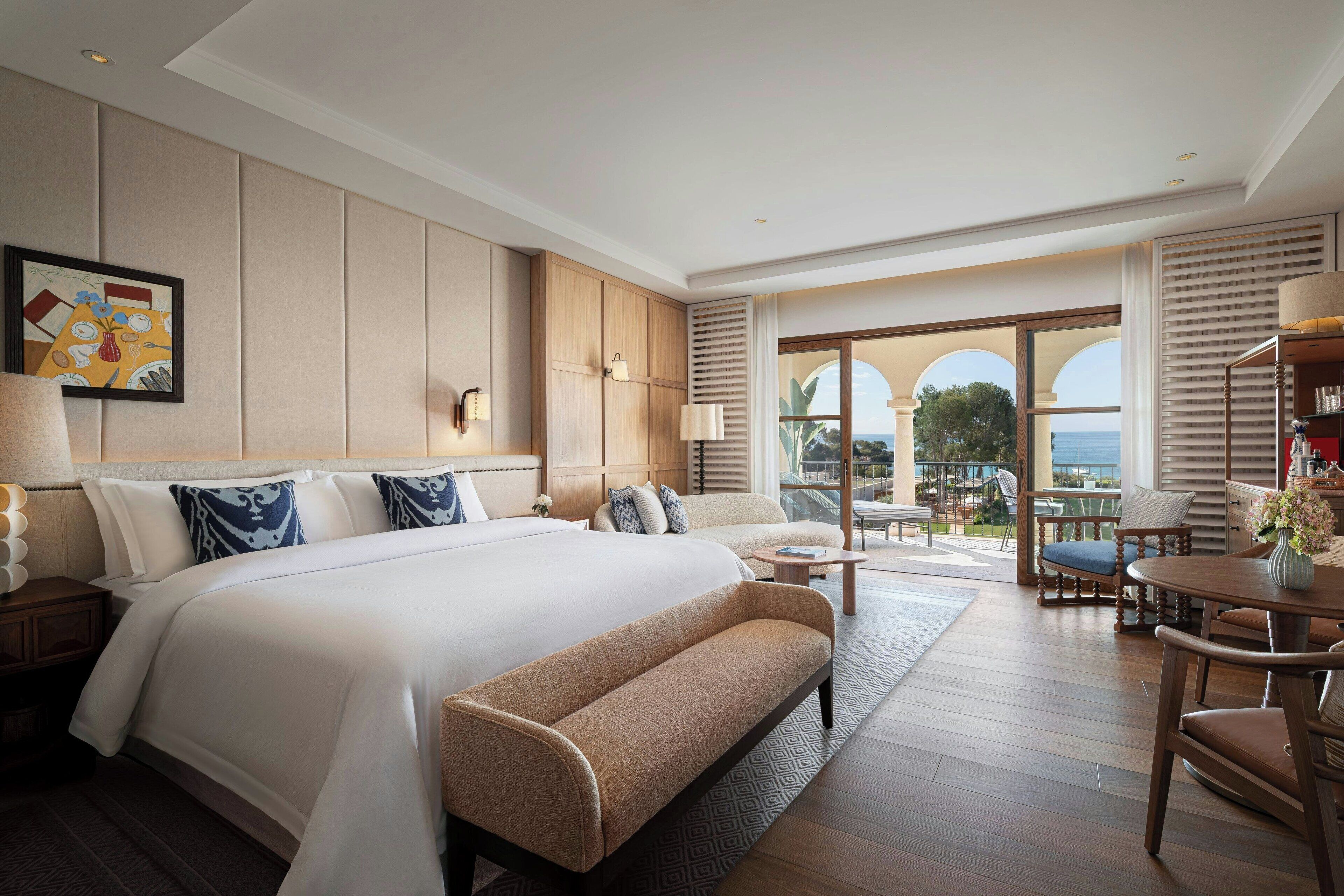 A spacious hotel room with a large bed, a sitting area with a bench, a dining table, and a view of the ocean through large windows.