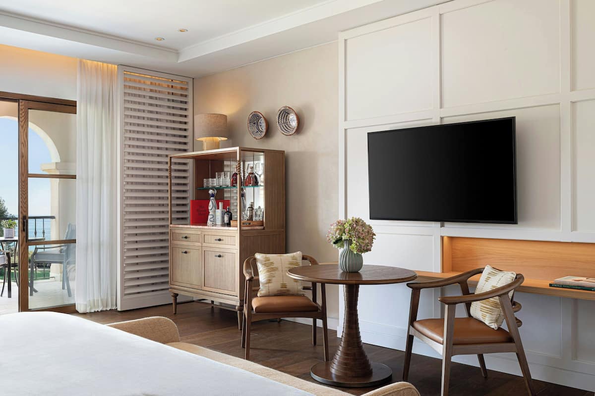 A hotel room with a large flat-screen TV, a wooden cabinet with glass doors, a round dining table, and a comfortable seating area.