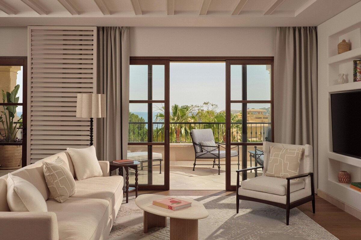 A modern living room with a sofa, armchair, and a coffee table. There is a balcony with a view of the sea and palm trees.