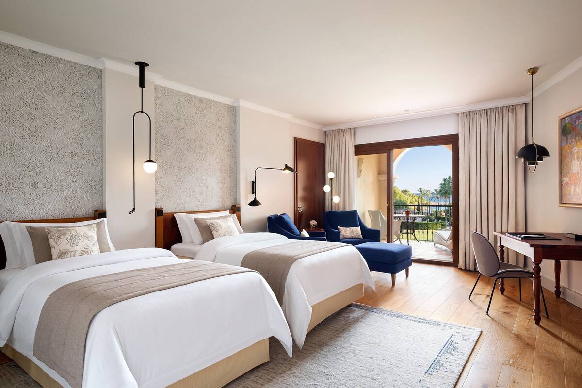 A hotel room with two beds, a desk, and a view of the outdoors.