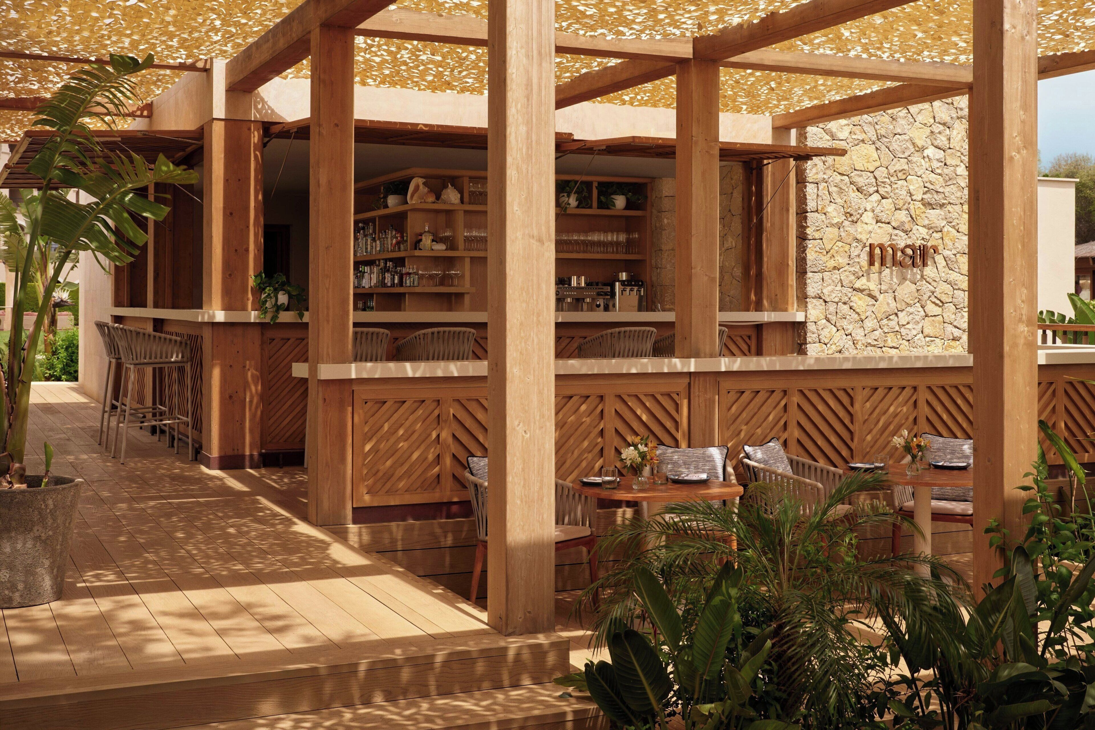 A wooden outdoor seating area with tables and chairs, surrounded by plants and a stone wall with the word 'moss' on it.