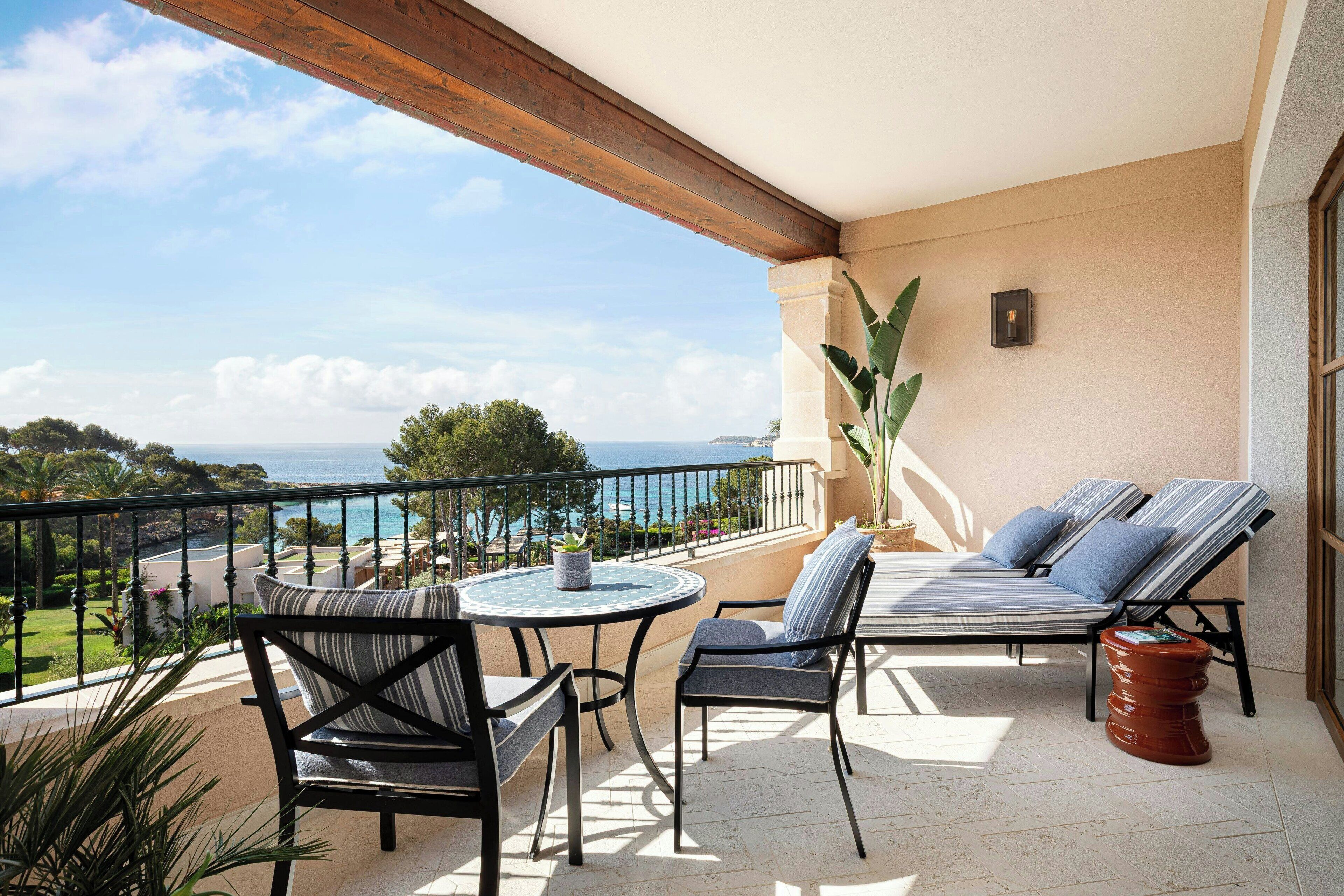 A balcony with a view of the sea, a round table with chairs, and a sofa with cushions.