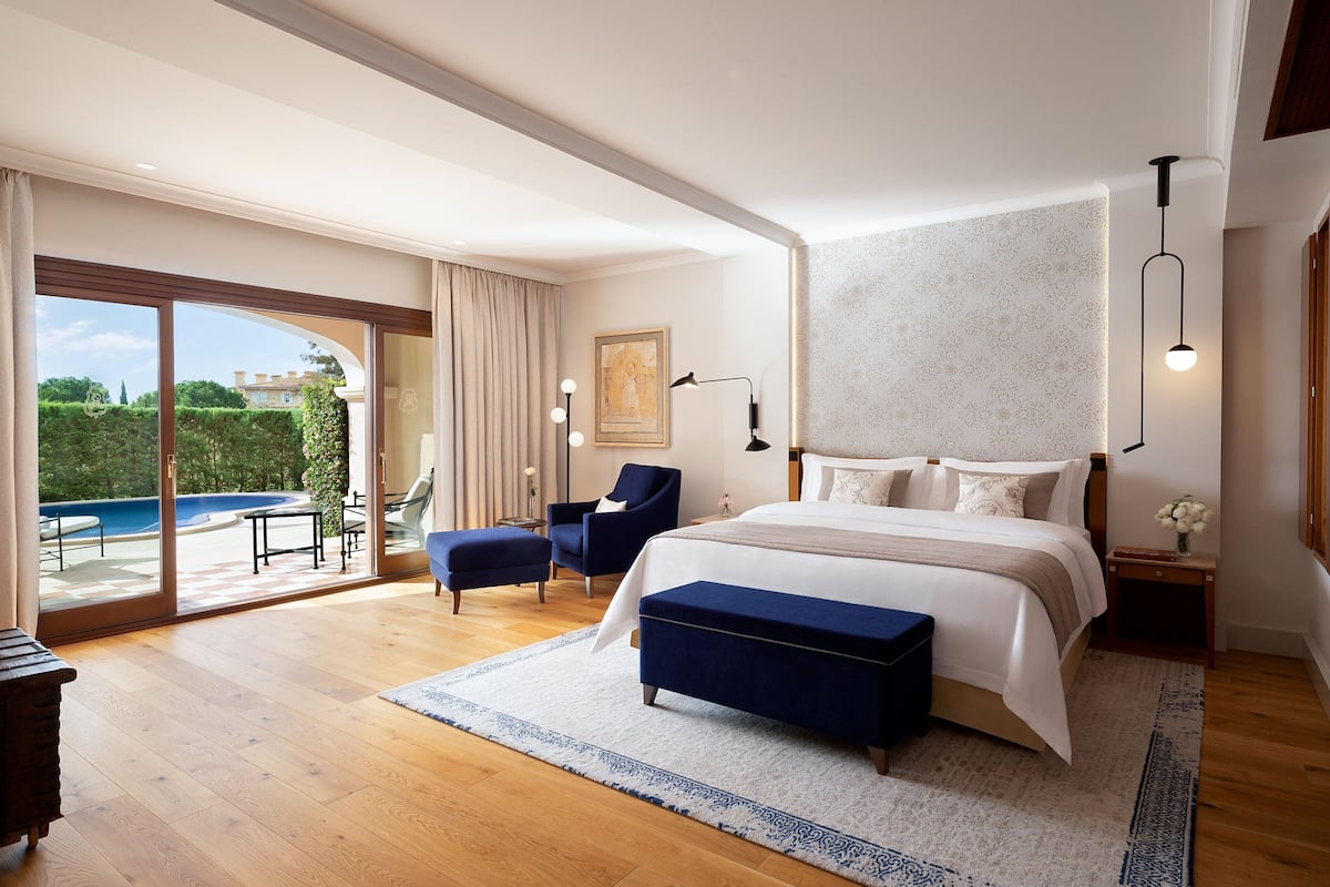 A spacious bedroom with a large bed, a seating area with a chair and ottoman, a desk, and a view of a swimming pool.