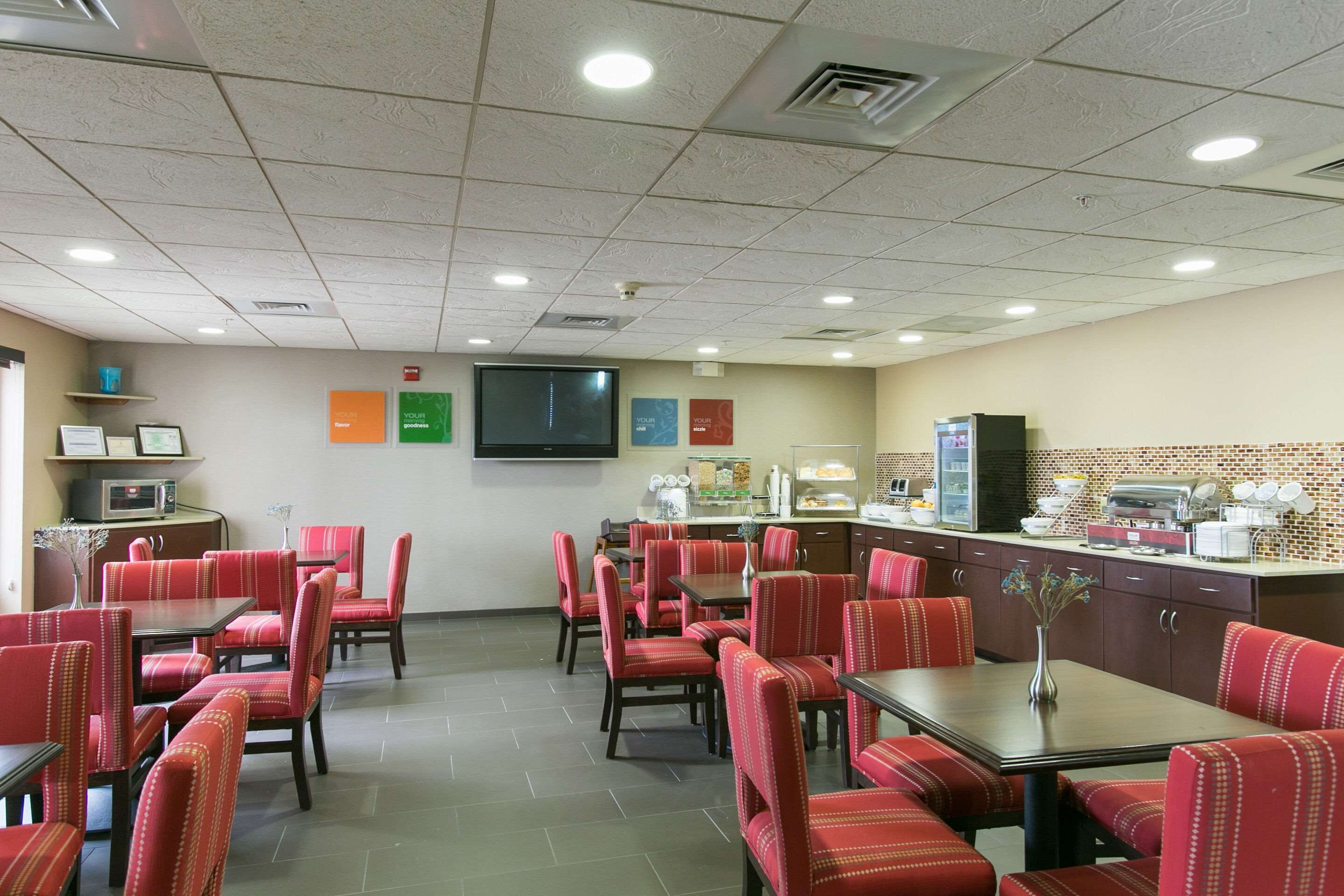 A dining area with red upholstered chairs and tables, a flat-screen TV mounted on the wall, and a kitchen area with a microwave and coffee maker.