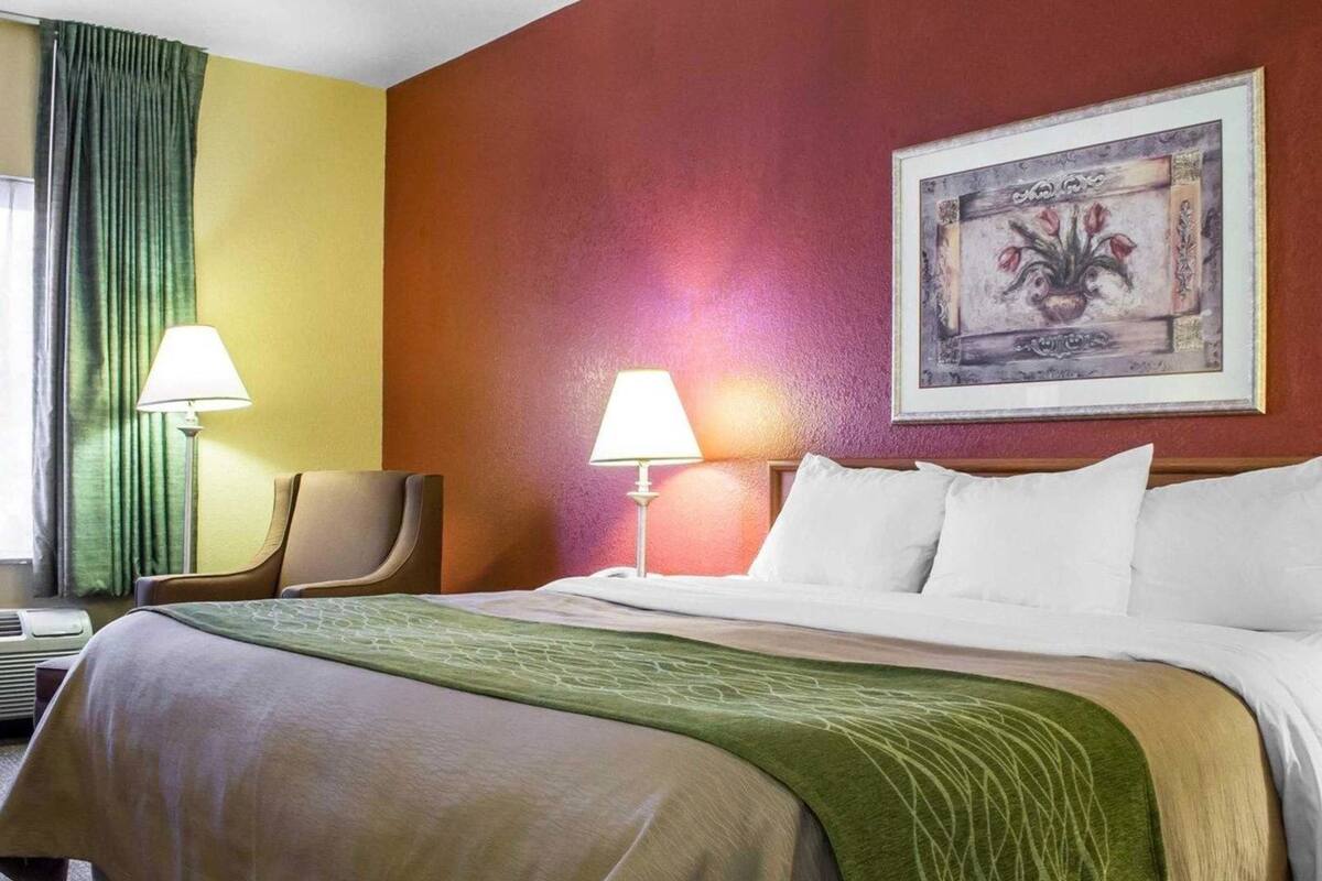 Ein Hotelzimmer mit einem Bett, zwei Nachttischlampen, einem Sessel, einem Bild an der Wand und einem grünen Vorhang.