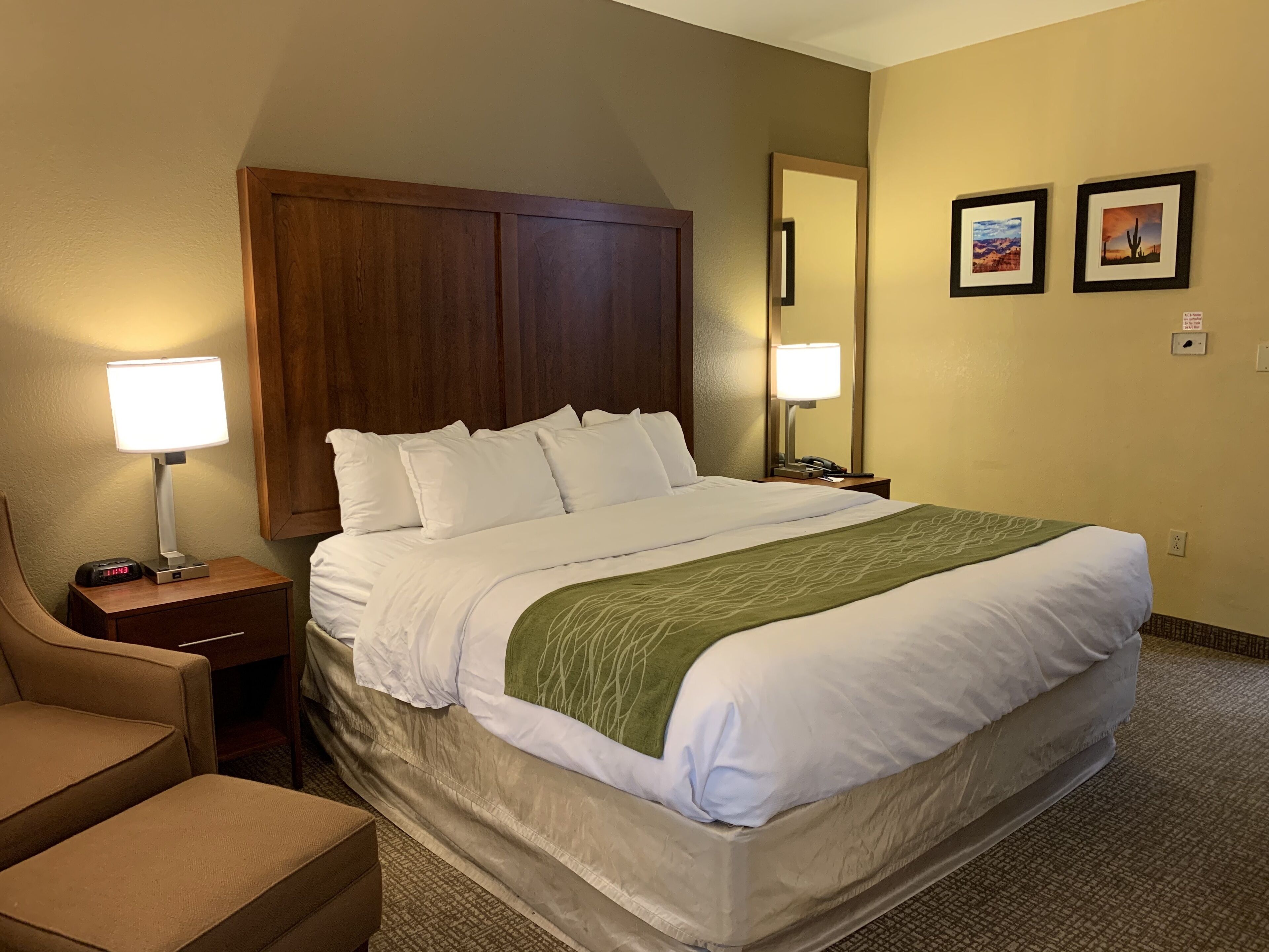 Habitación de hotel con una cama grande, cabecera de madera, mesita de noche con lámpara, una silla y dos cuadros en la pared.