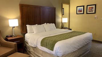 Habitación de hotel con una cama grande, cabecera de madera, mesita de noche con lámpara, una silla y dos cuadros en la pared.