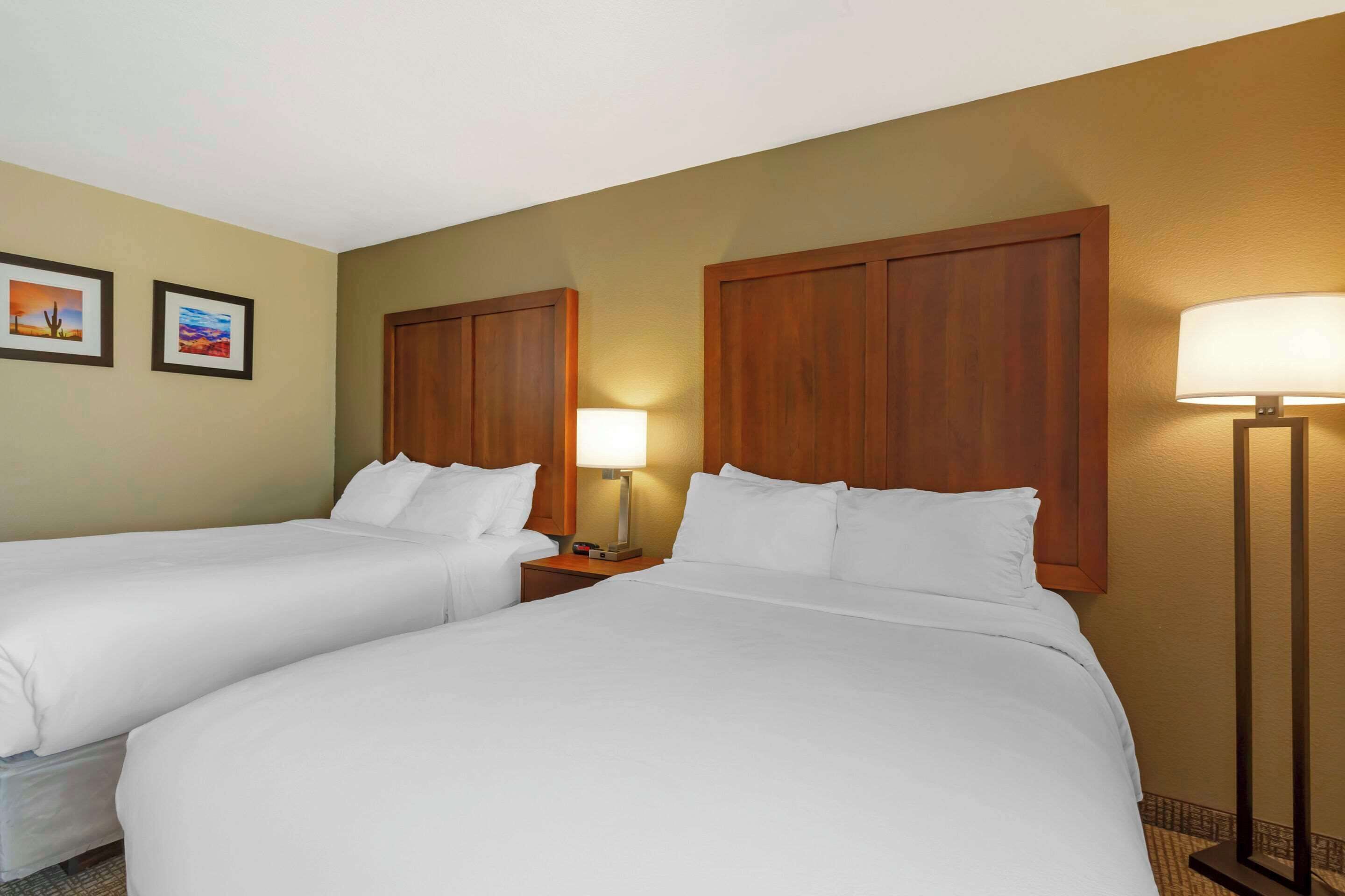 A hotel room with two beds, wooden headboards, a nightstand with a lamp, and two framed pictures on the wall.