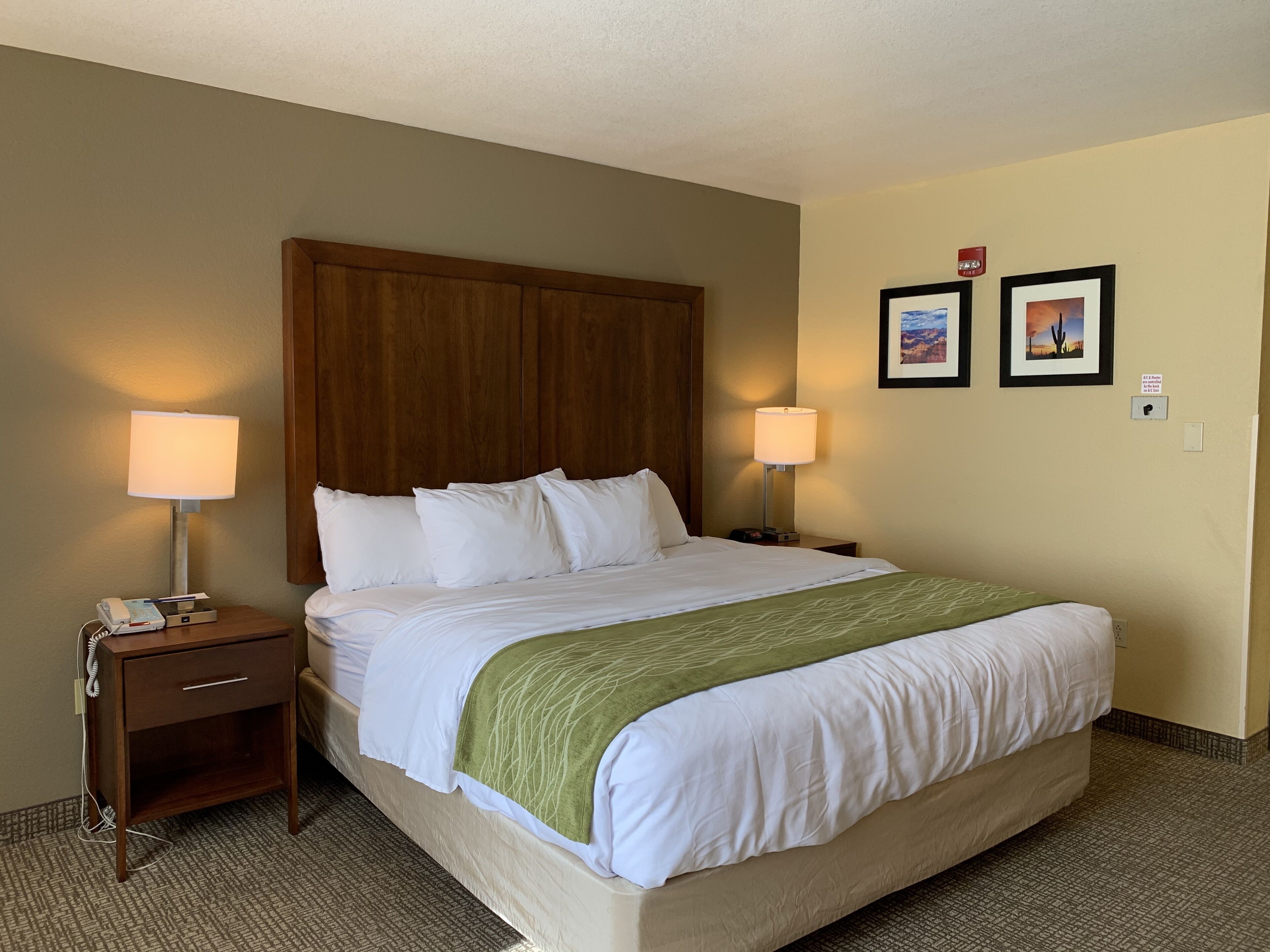 A hotel room with a large bed, two bedside lamps, a wooden headboard, a green throw blanket, and two framed pictures on the wall.