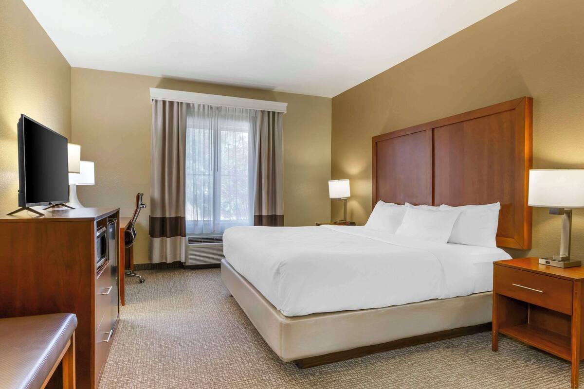 Ein Hotelzimmer mit einem großen Bett, einem Schreibtisch mit Fernseher, einer Lampe und einem Vorhangfenster.