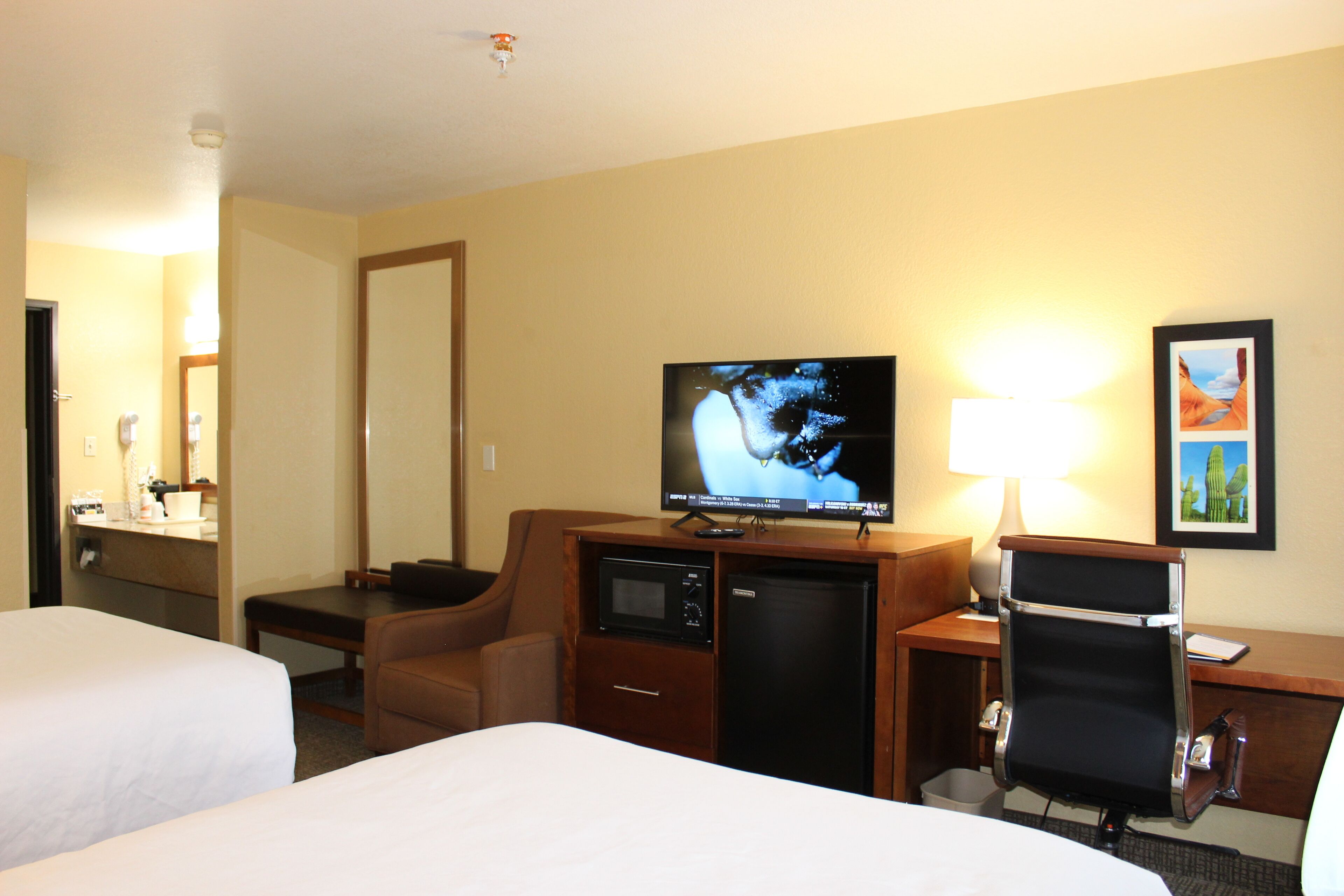 Quarto de hotel com duas camas, uma mesa de trabalho com televisão, micro-ondas, frigorífico, uma cadeira e abajur.