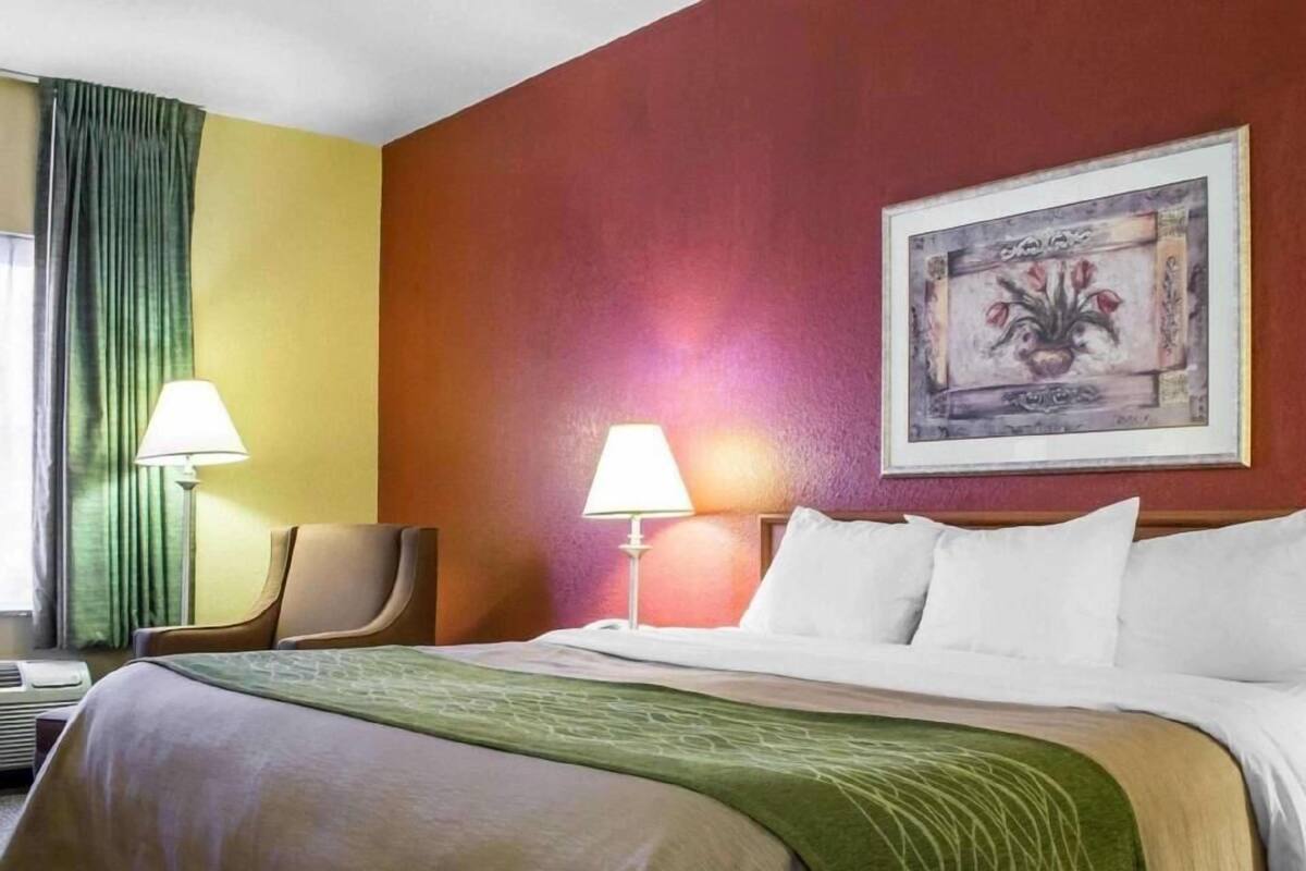 Ein Hotelzimmer mit einem Bett, zwei Nachttischlampen, einem Sessel, einem Wandbild und einem Fenster mit Vorhängen.