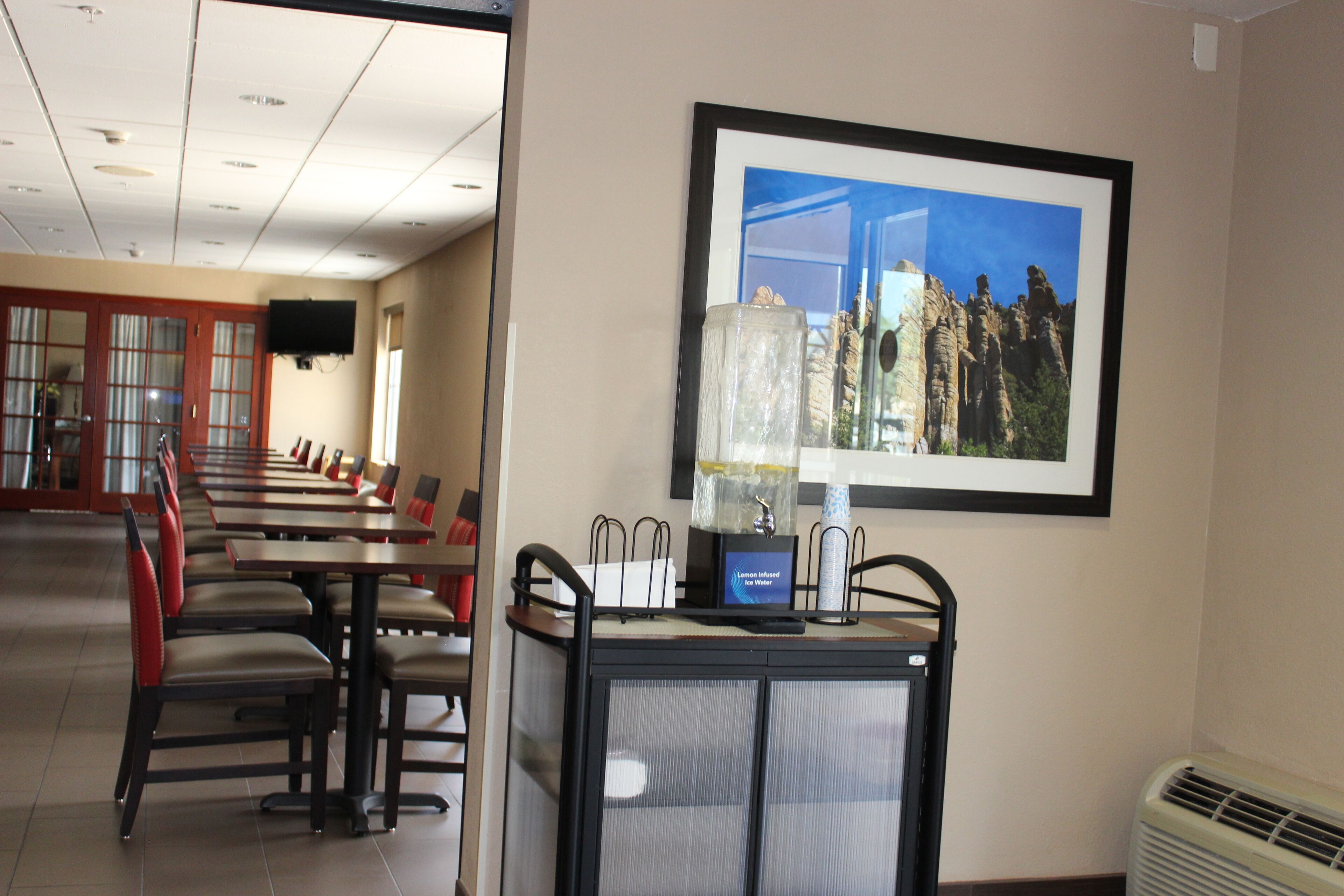 A hotel dining area with tables and chairs, a framed picture on the wall, and a small kiosk with a display.