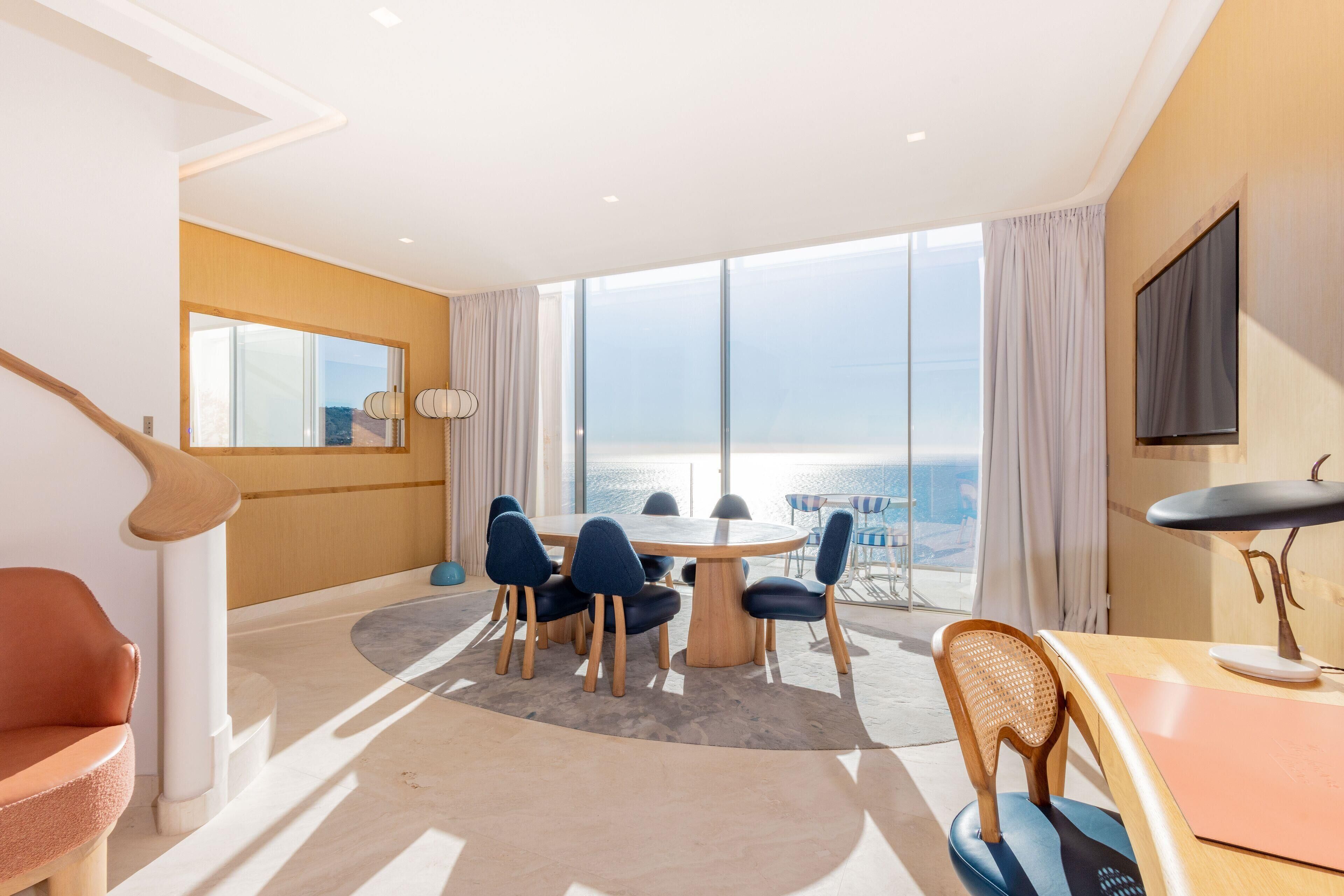 A modern hotel room with a round dining table, multiple chairs, a wooden desk, a TV, and a large window with a view of the sea.
