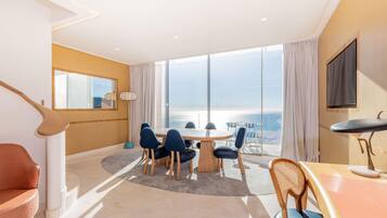 A modern hotel room with a round dining table, multiple chairs, a wooden desk, a TV, and a large window with a view of the sea.