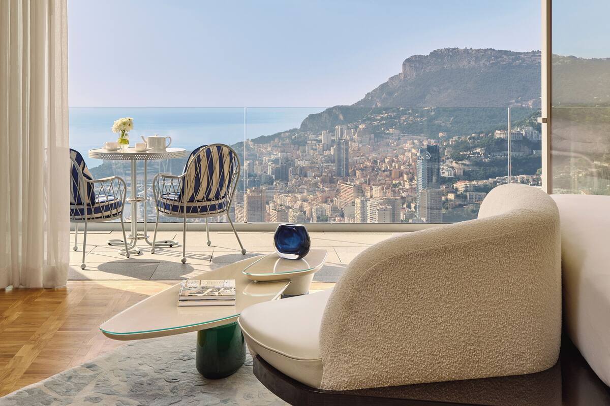 A modern living room with a sofa, chairs, and a small table with a vase and teapot. A large window offers a view of a cityscape and mountains.
