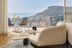 A modern living room with a sofa, chairs, and a small table with a vase and teapot. A large window offers a view of a cityscape and mountains.