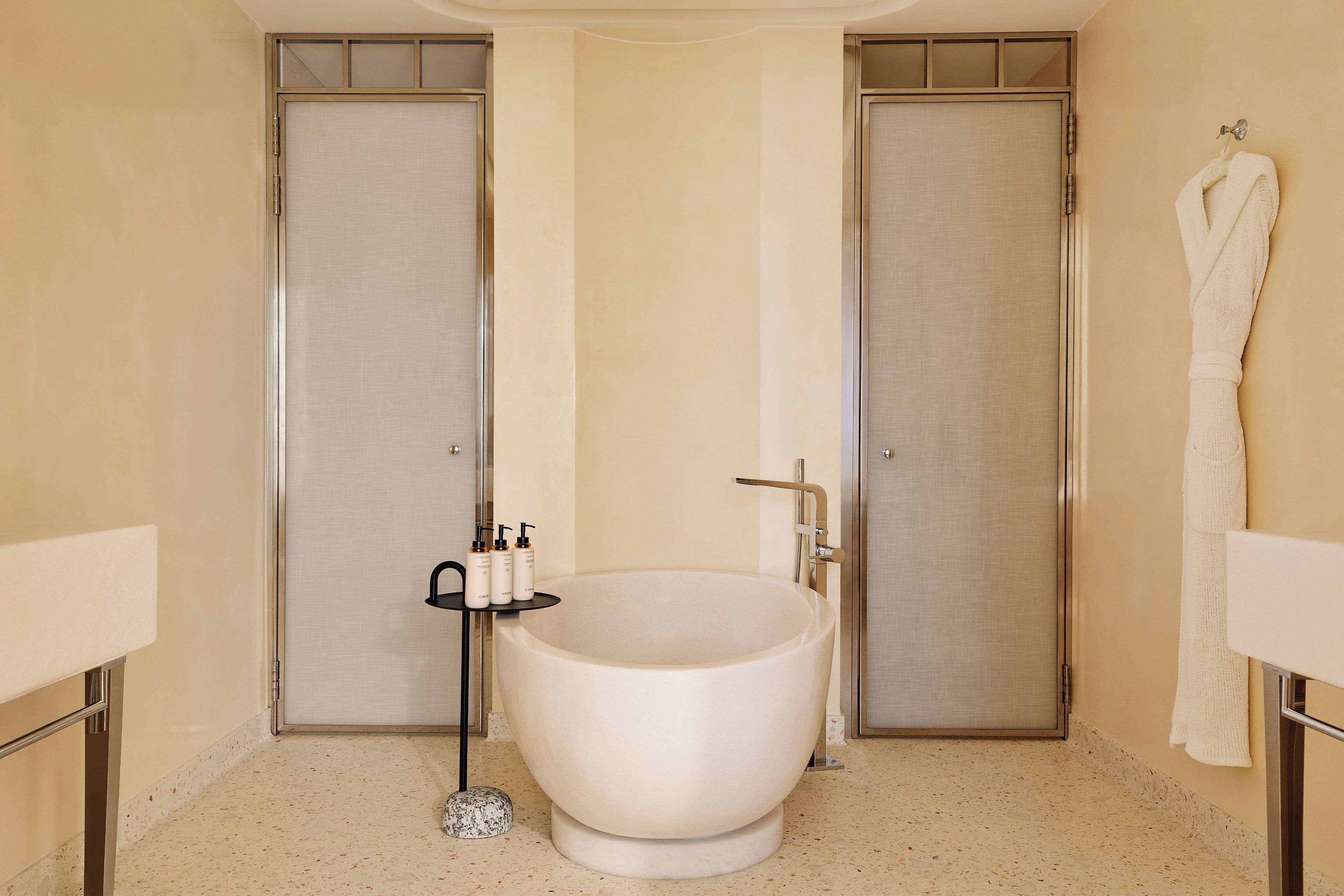 A bathroom with a freestanding bathtub, two doors, a towel rack, and a small table with toiletries.