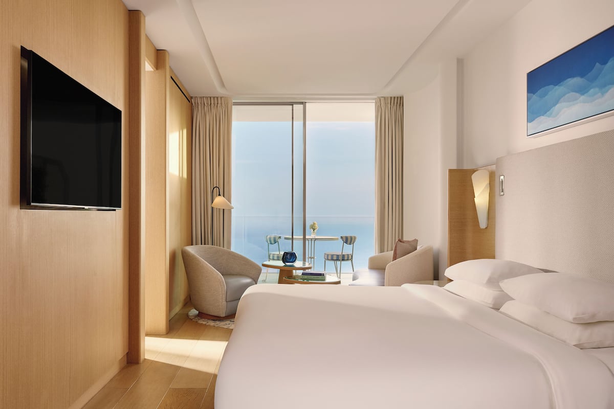 A hotel room with a large bed, a TV, a balcony with a table and chairs, and a view of the ocean.