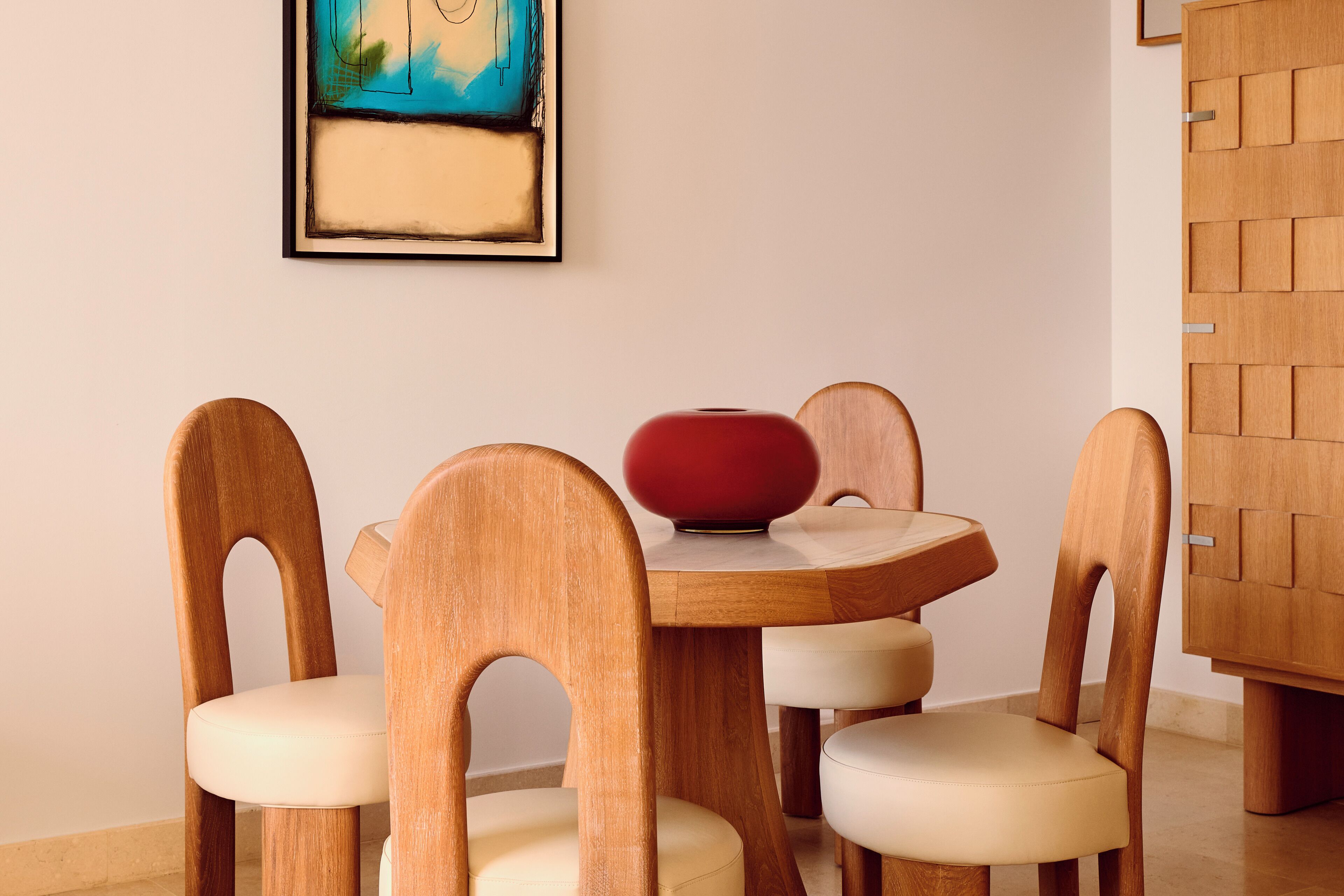 Un comedor con una mesa redonda de madera, cuatro sillas de madera con asientos acolchados y un florero rojo sobre la mesa.