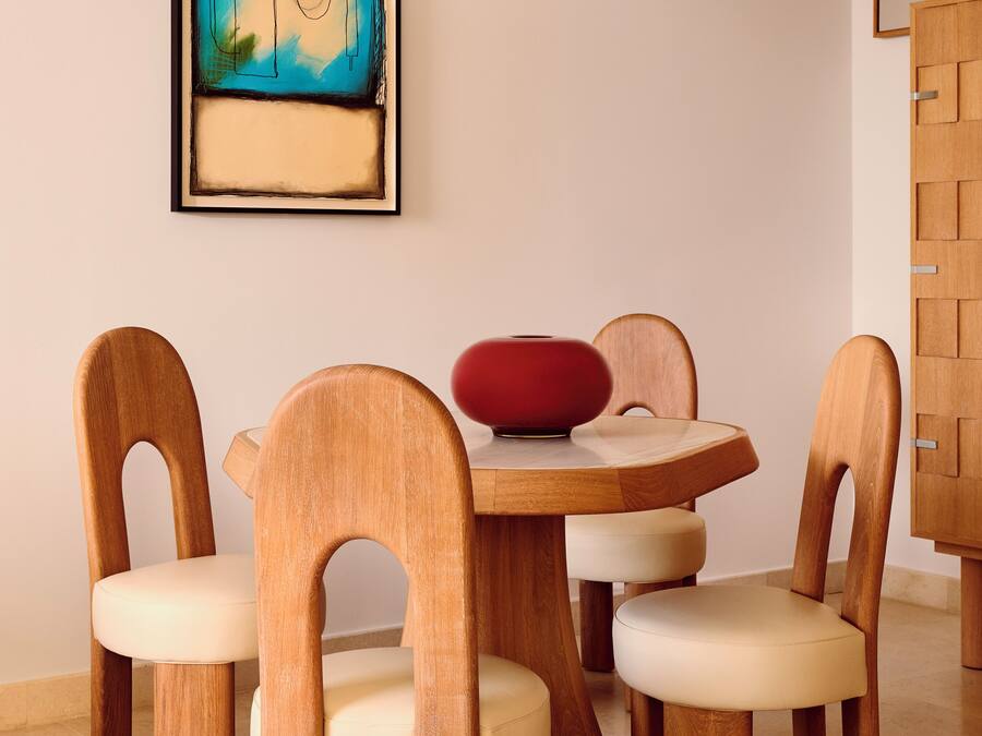 Un comedor con una mesa redonda de madera, cuatro sillas de madera con asientos acolchados y un florero rojo sobre la mesa.