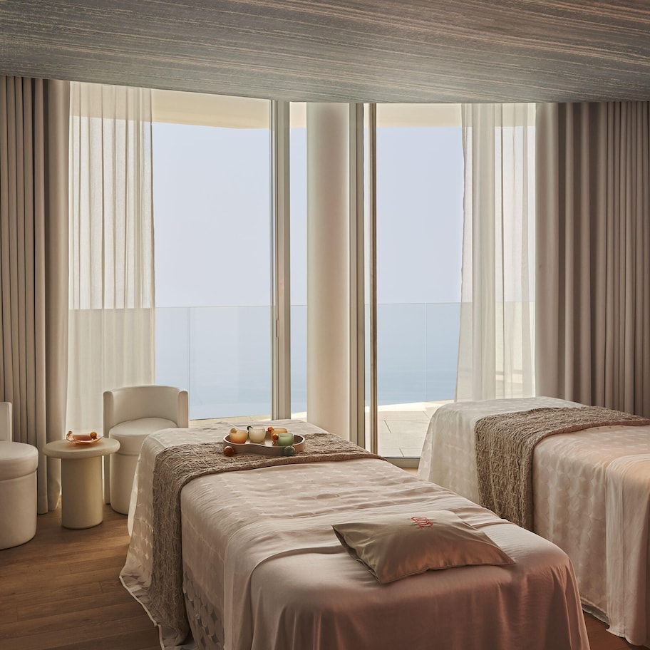 A hotel room with two beds, a sofa, a small table with a tray of fruit, and large windows with curtains.