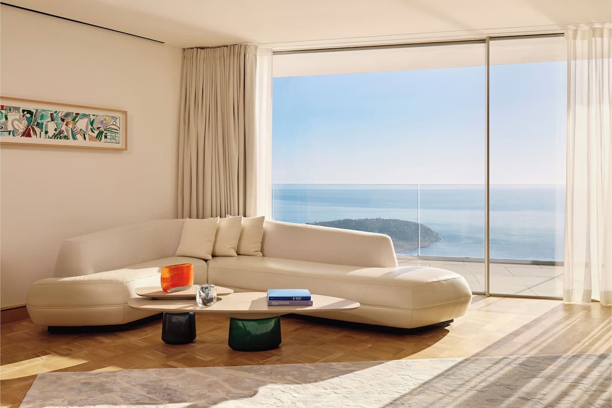 A modern living room with a large sectional sofa, a coffee table, and a view of the ocean through floor-to-ceiling windows.