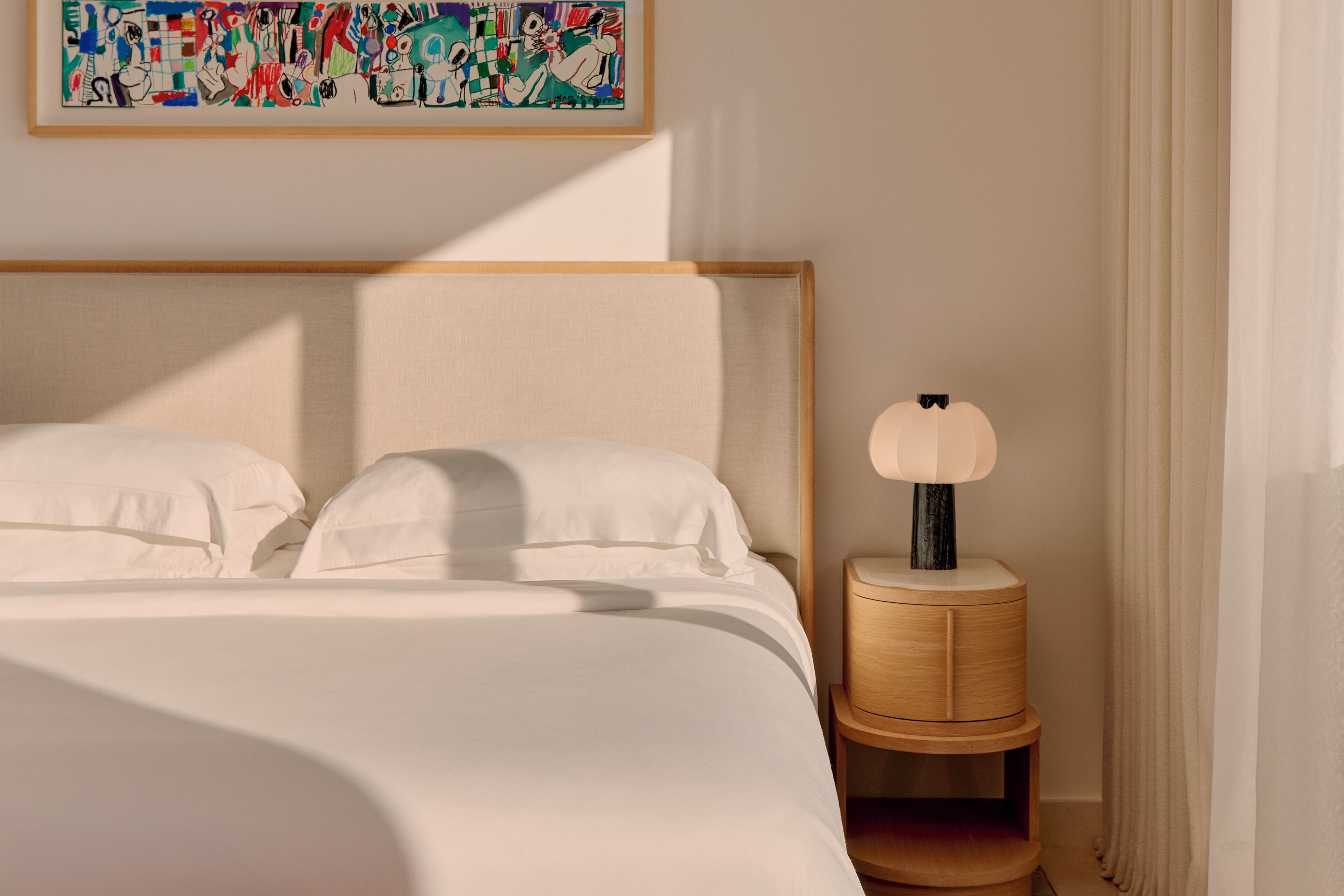 A neatly made bed with white linens, a wooden headboard, a bedside table with a lamp, and a colorful abstract painting on the wall.