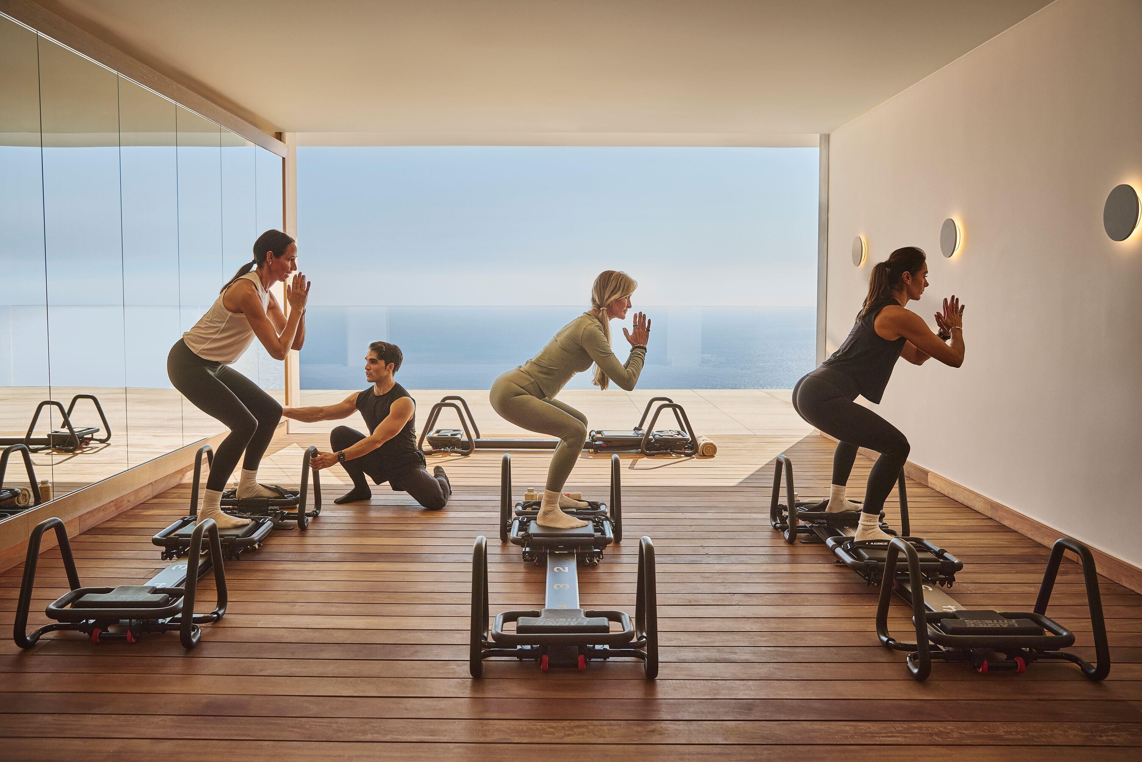 A modern fitness studio with large windows and a wooden floor, featuring exercise equipment and mirrors.