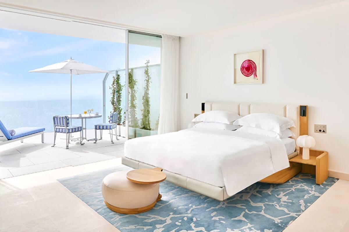 A bedroom with a large bed, a small round ottoman, a bedside table with a lamp, a chair, and a balcony with a table and chairs.
