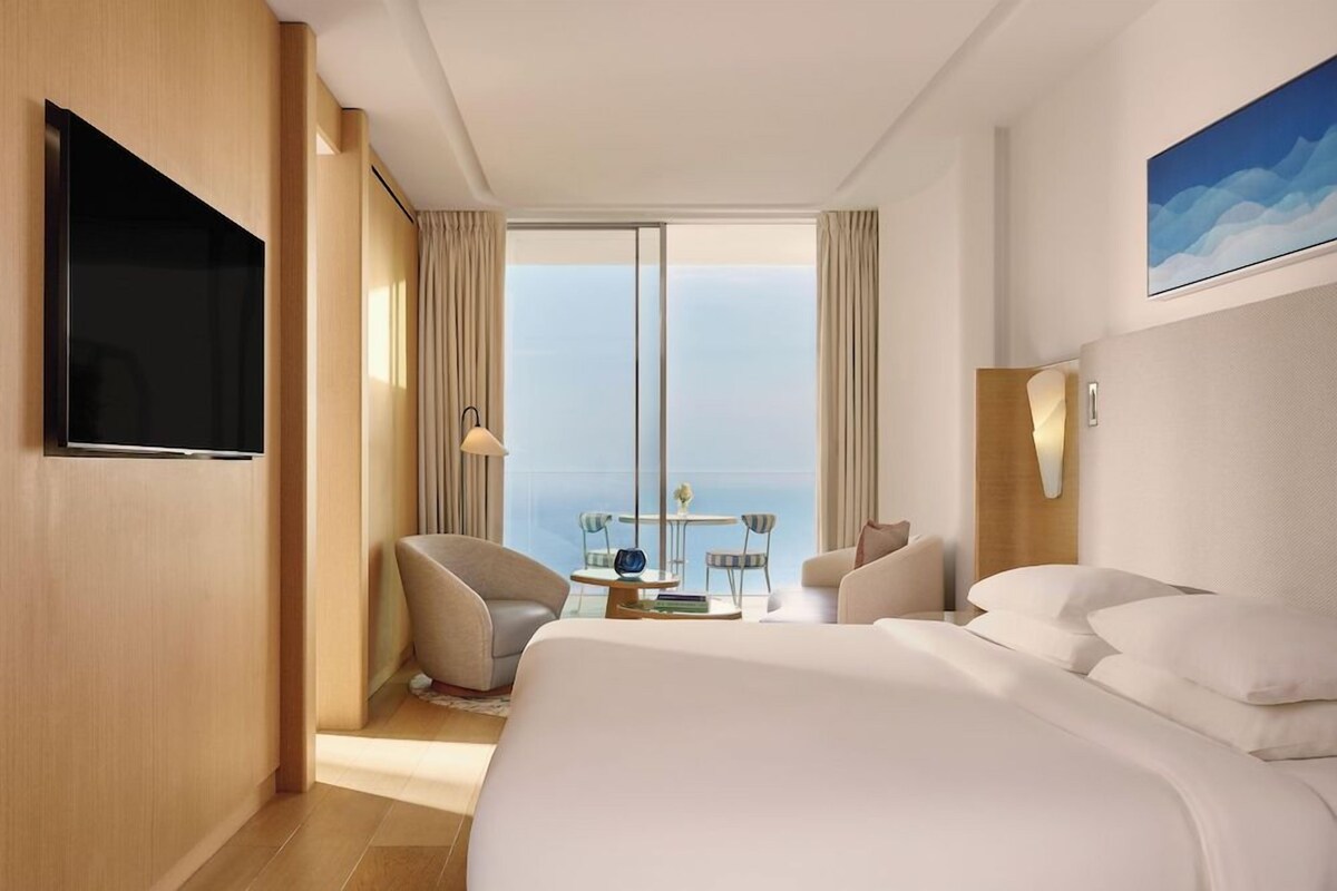 A hotel room with a large bed, a TV, a balcony with a view, and a small table with chairs.