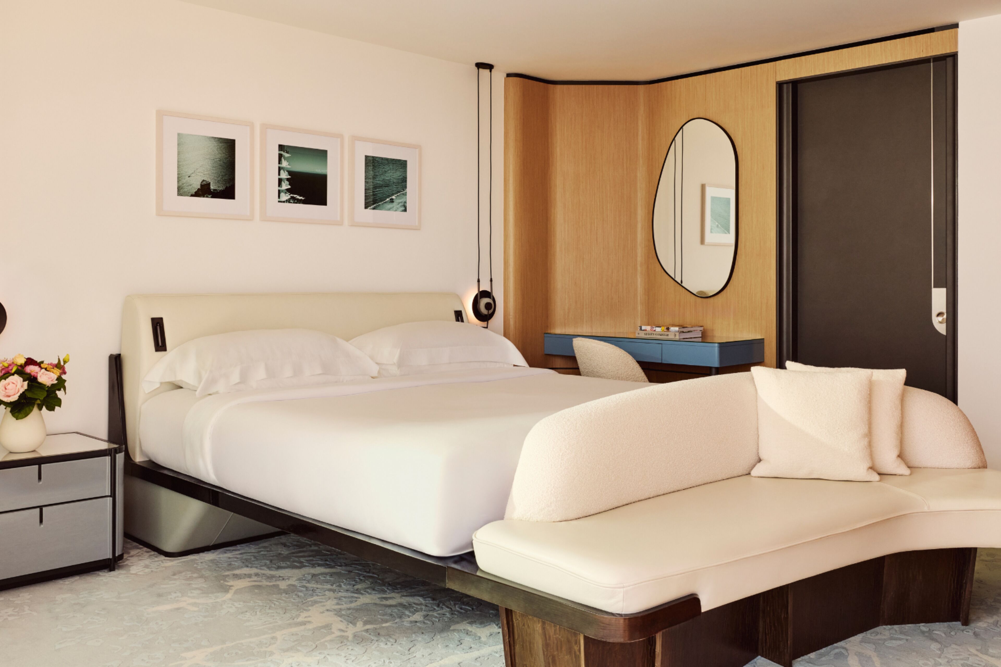 Un dormitorio moderno con una cama grande, una mesita de noche con un jarrón de flores, un banco empotrado, un espejo y cuadros enmarcados en la pared.