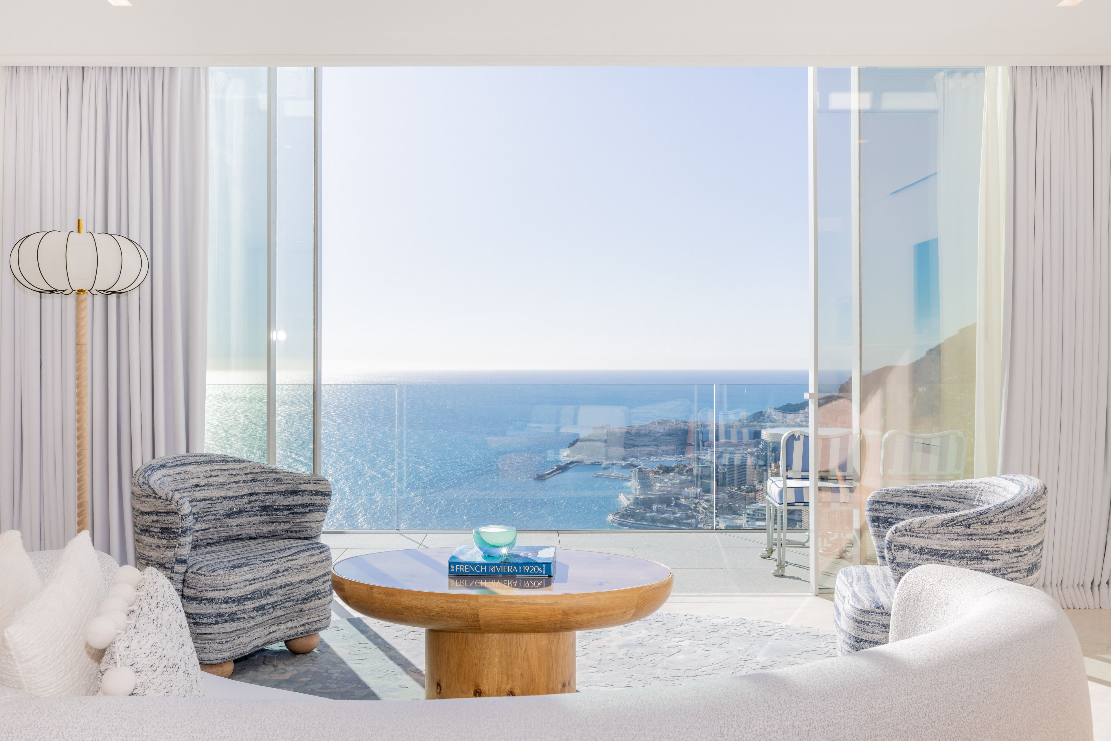 A modern living room with a large window, two armchairs, a round wooden coffee table, and a view of the sea.