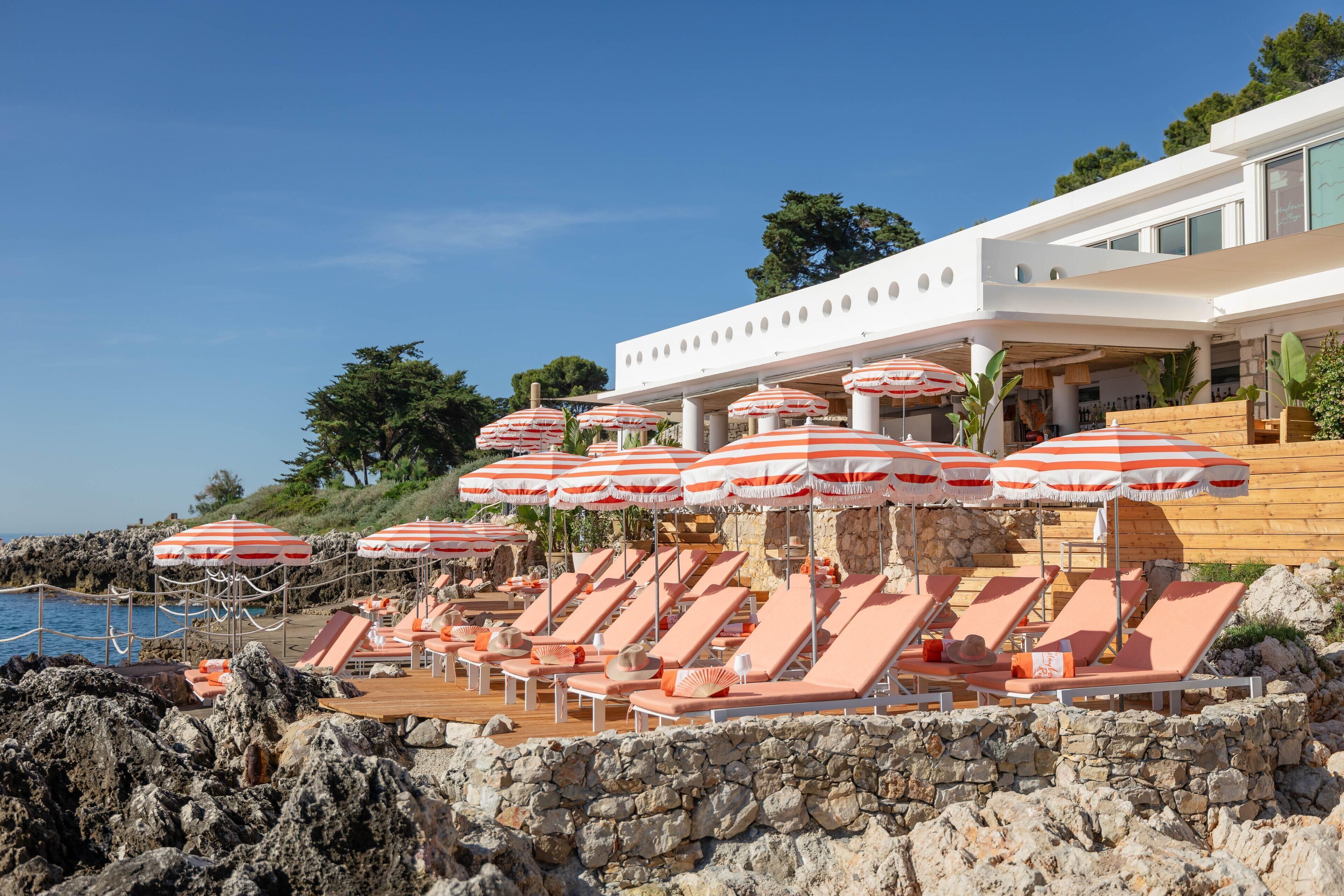 Un espace aménagé au bord de la piscine, avec des chaises longues et des parasols à rayures, un mur en pierre et un bâtiment moderne en arrière-plan.