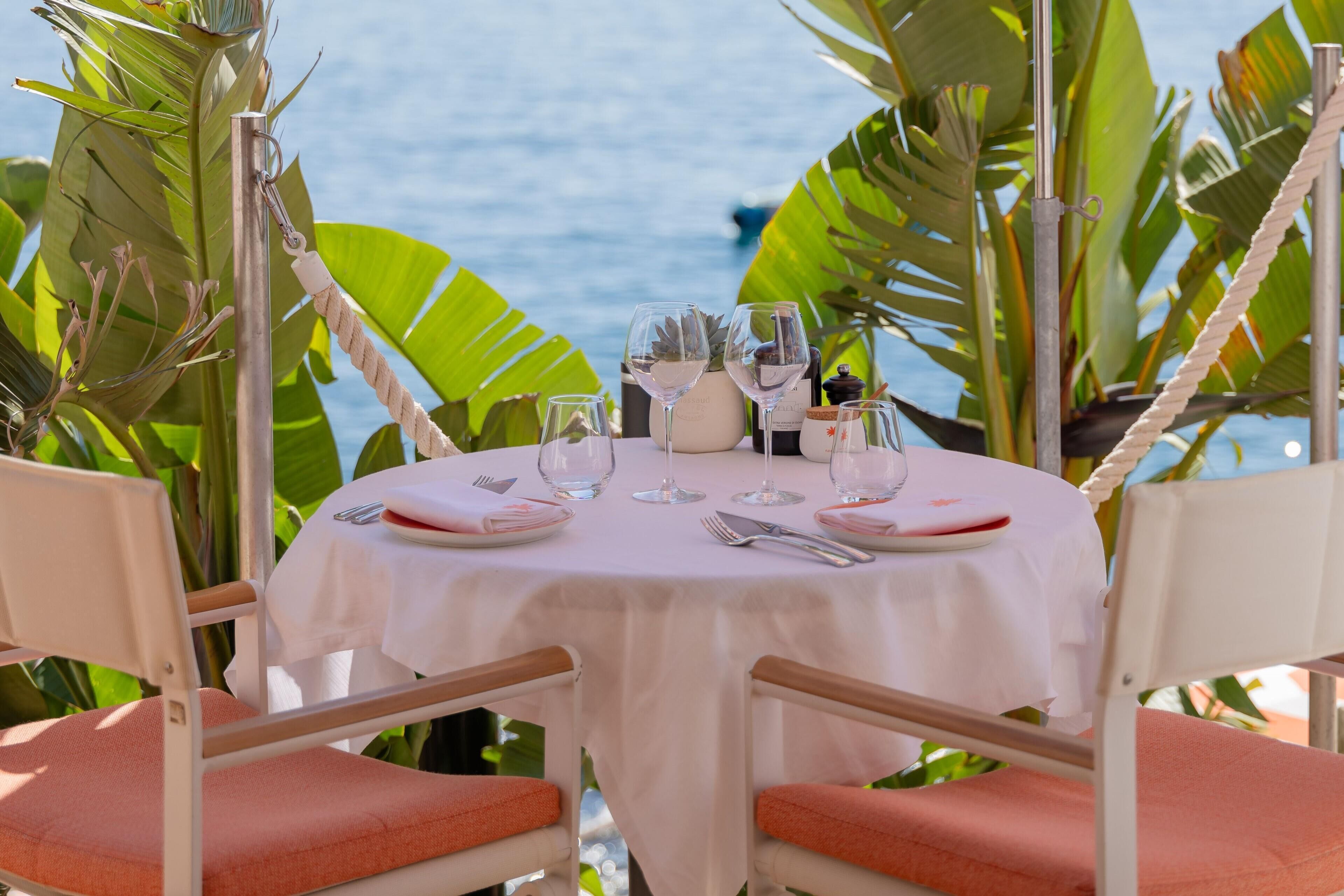 Une table ronde recouverte d’une nappe blanche, entourée de chaises dotées de coussins orange, dressée pour un repas avec des verres à vin, des assiettes et des couverts.
