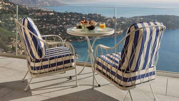 Two striped chairs with a table and a bowl of fruit and juice on a balcony overlooking a coastal view.