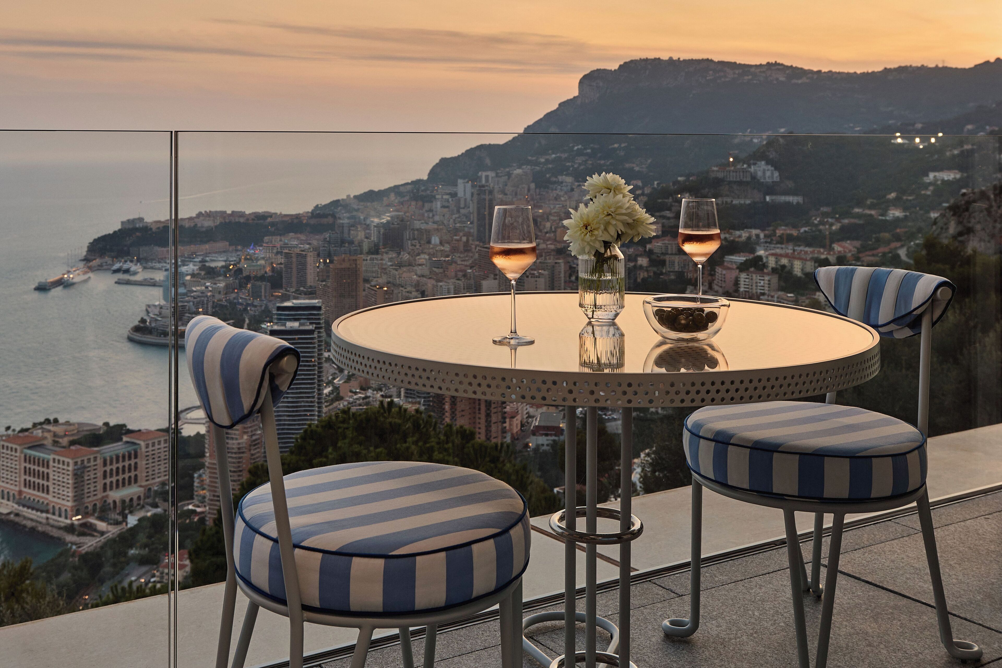 Un tavolo rotondo con due sedie, un vaso con fiori e due bicchieri da vino su un balcone con vista sulla città e sul mare.