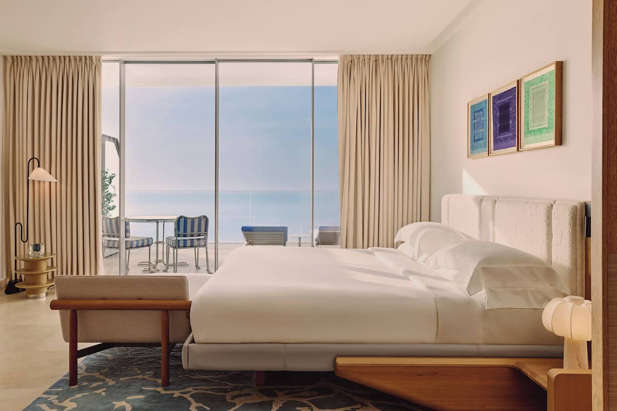 A hotel room with a large bed, a small table, a chair, and a view of the sea through a sliding door.