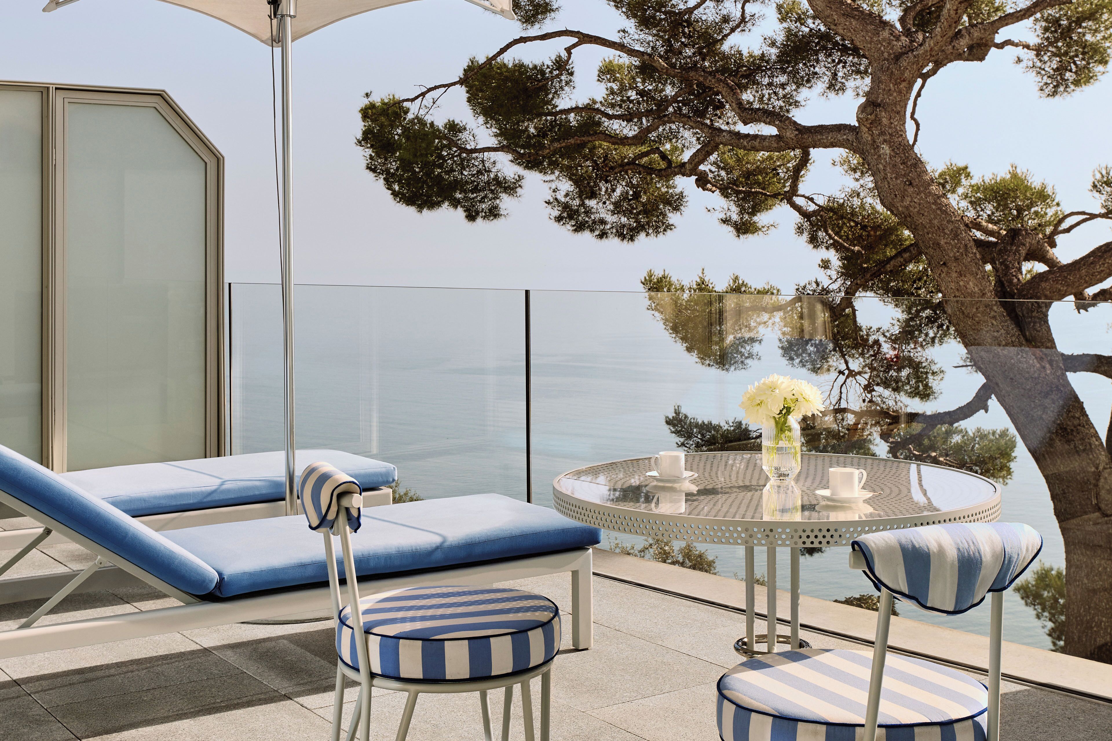 Un balcon avec des fauteuils de jardin bleus, une table ronde en verre surmontée d’un vase blanc, et une vue sur la mer.