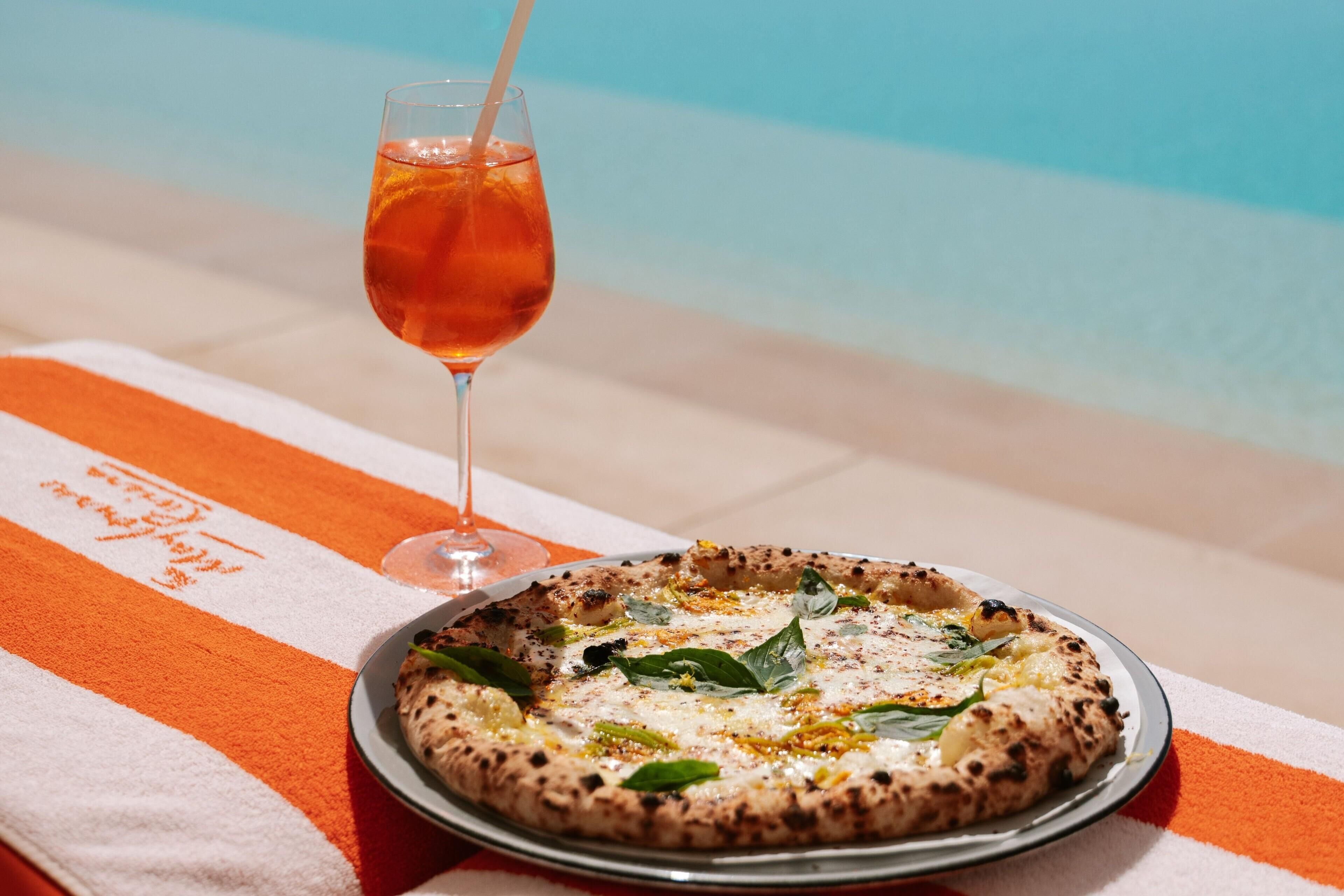 A pizza on a plate with a glass of drink on a towel by a pool.