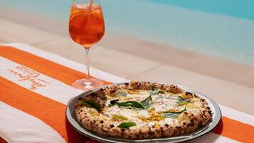 A pizza on a plate with a glass of drink on a towel by a pool.