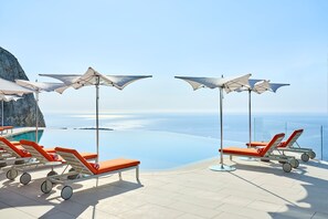 A poolside area with sun loungers and umbrellas, overlooking a clear blue sea and a rocky coastline.